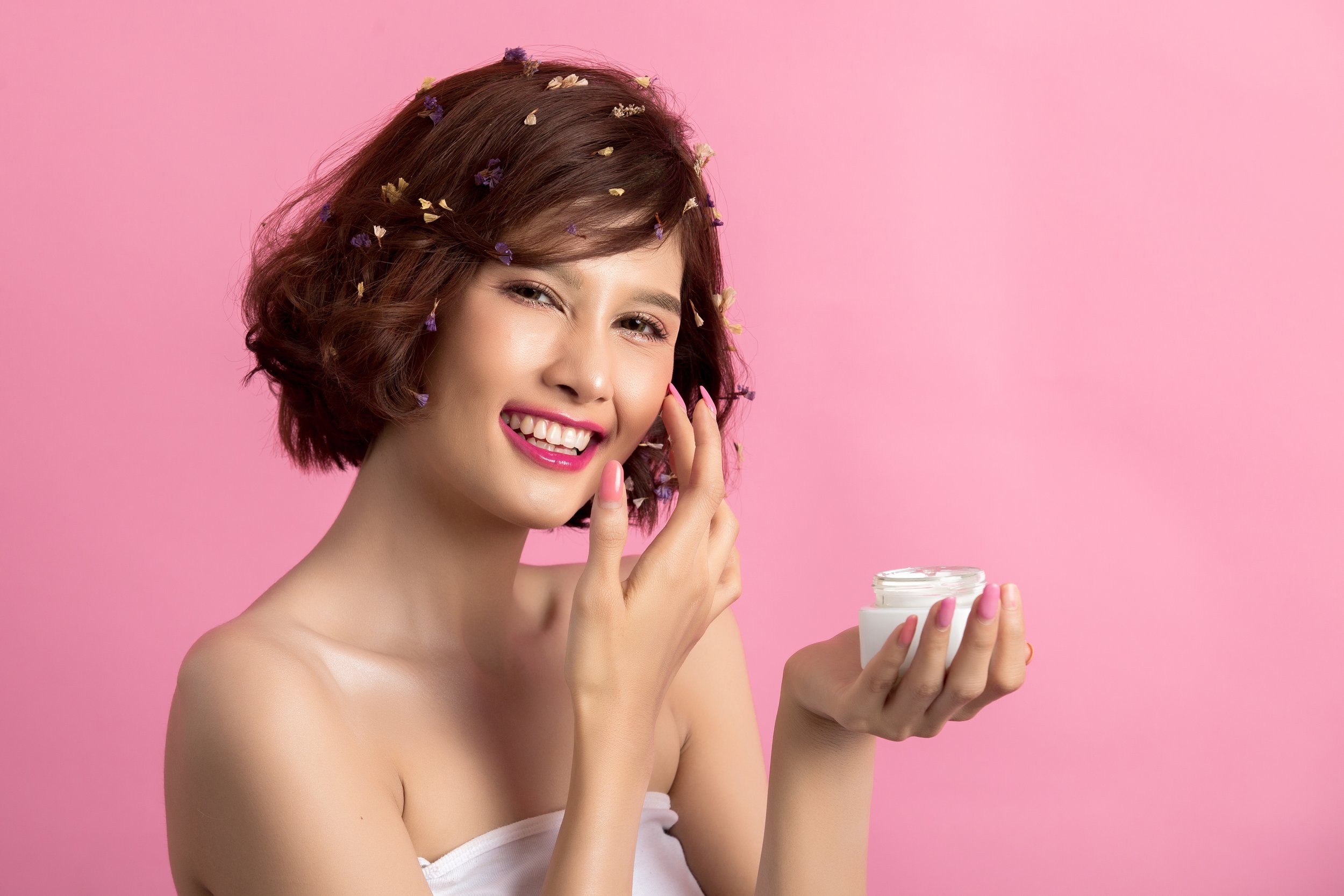 A young woman with short brown hair adorned with small flowers, smiling and holding a jar of skincare cream against a pink background.