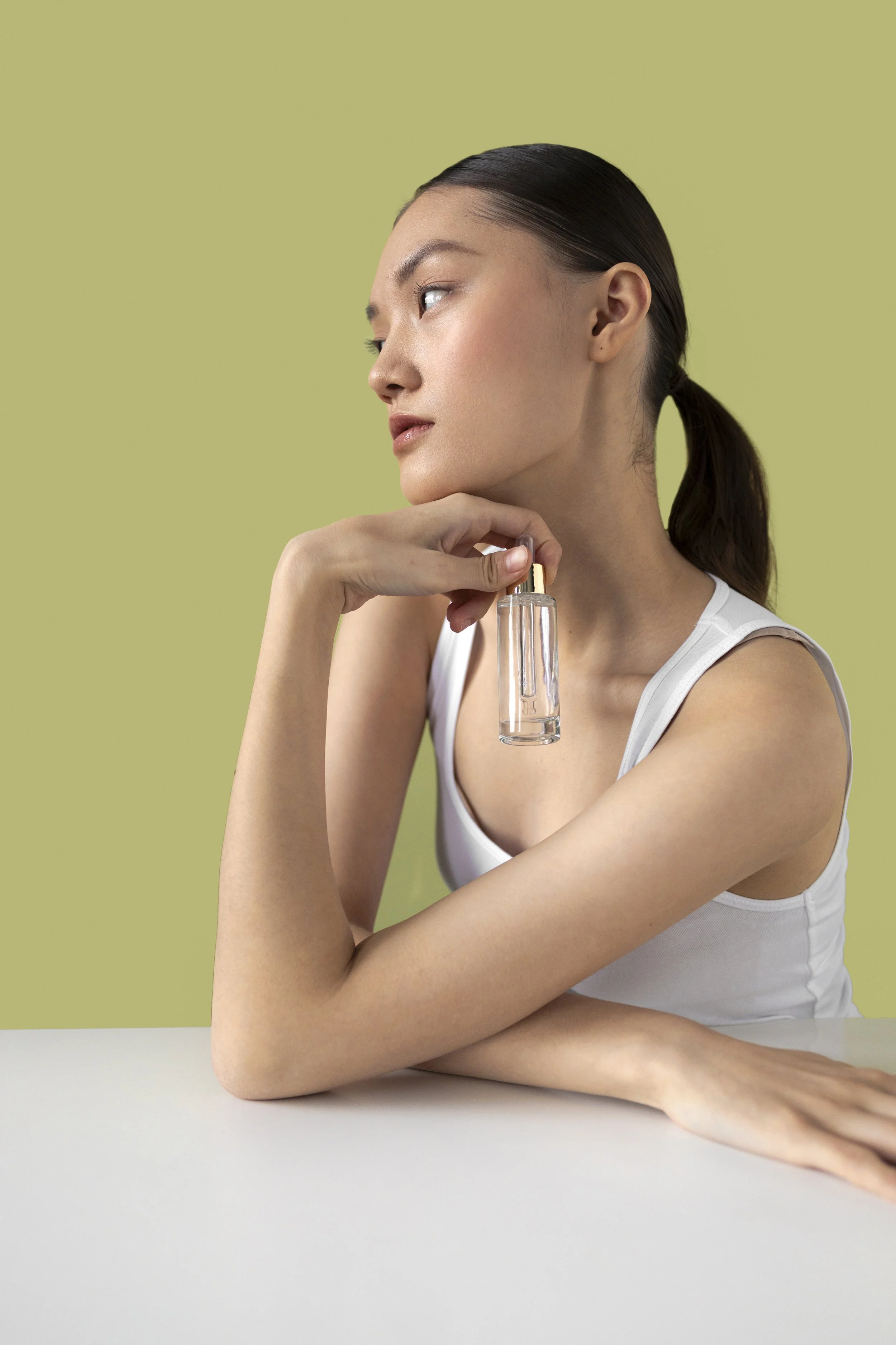 A woman with dark hair tied back in a ponytail, wearing a white sleeveless top, holding a small glass spray bottle near her neck, with a contemplative expression against a light green background.