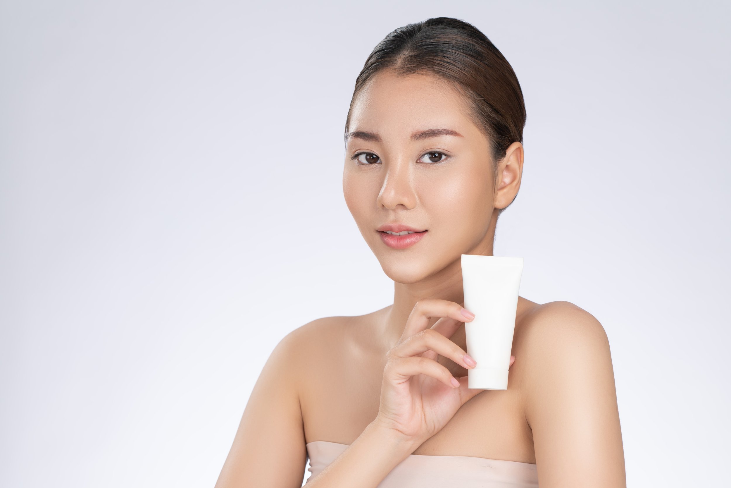 A woman with smooth skin holding a white cosmetic tube in front of her, smiling softly, against a plain light background.