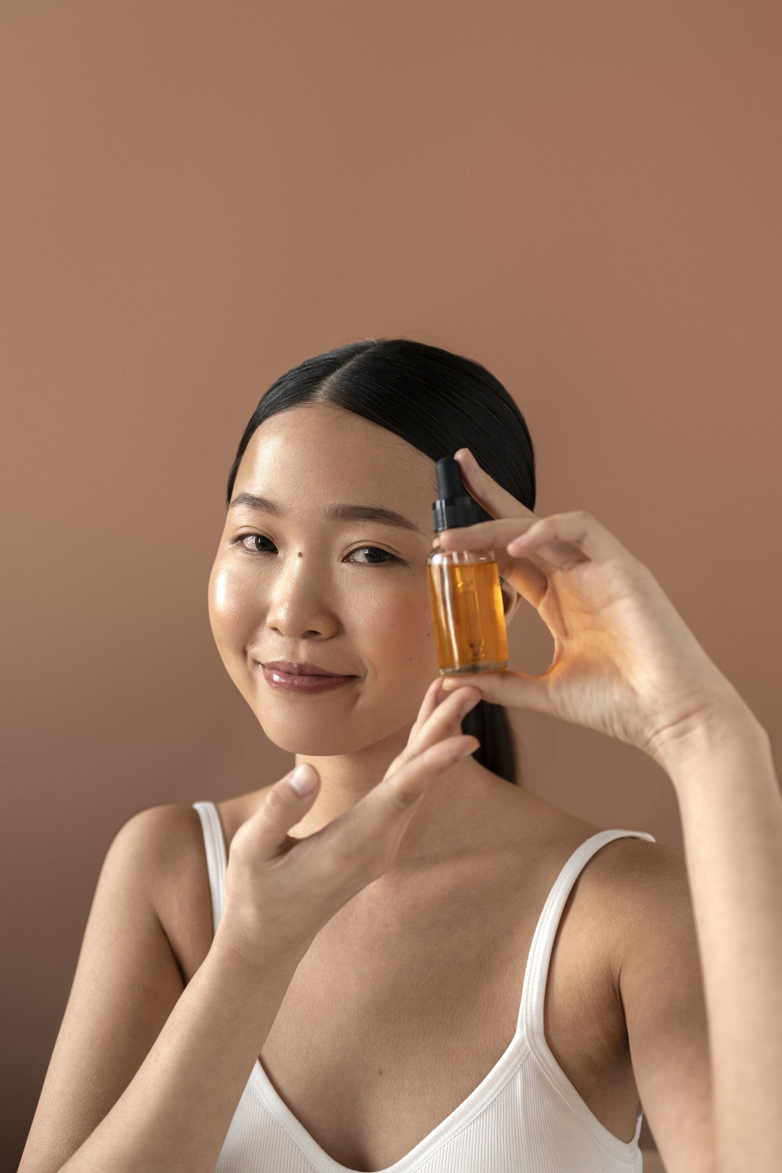 A woman holding a small amber glass bottle with a dropper cap, smiling softly against a neutral background.