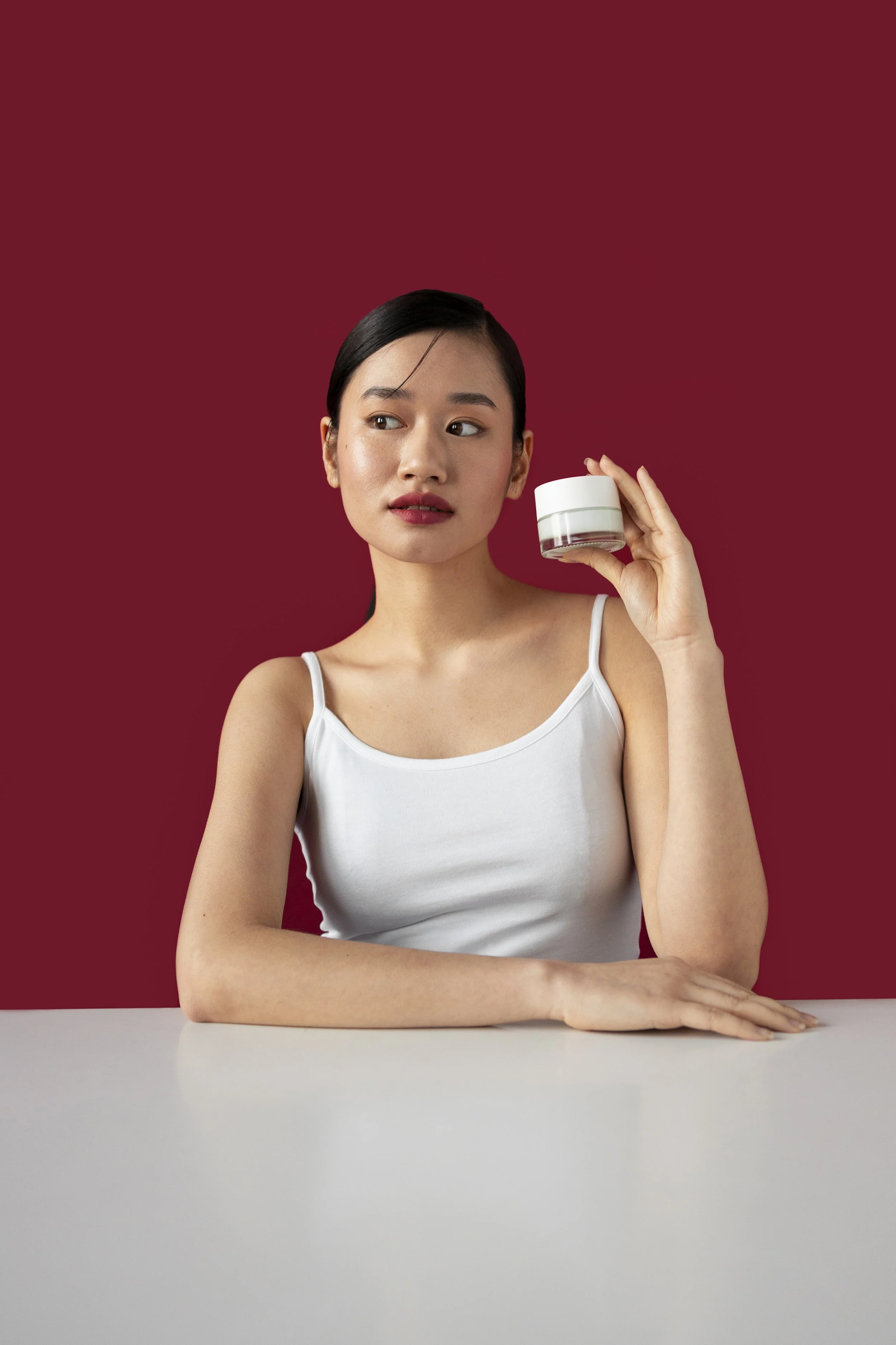 A young woman with dark hair and a white tank top holding a small white jar, sitting at a white table against a red background.