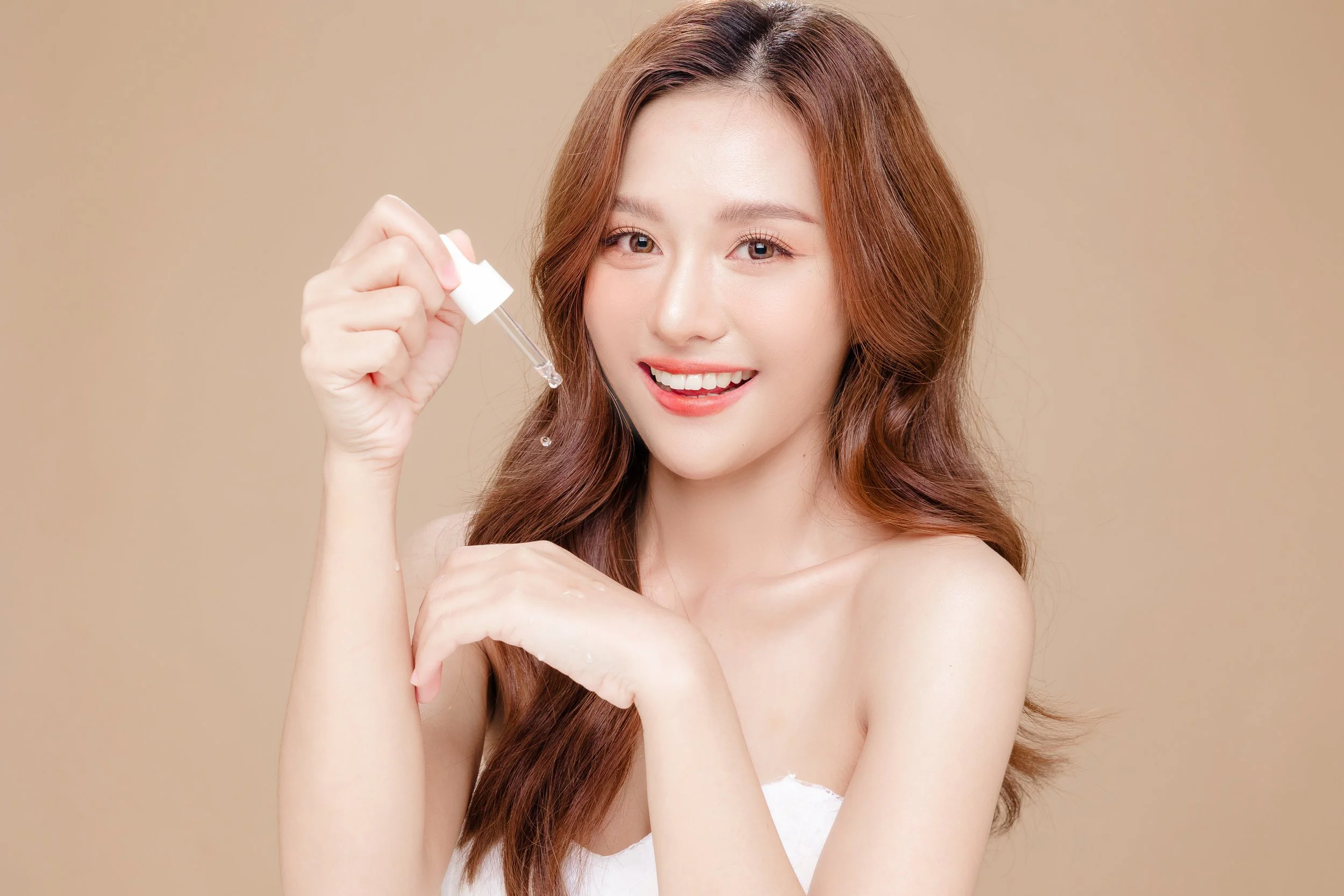 Young woman with long wavy hair holding a dropper and applying skincare product, smiling at the camera.