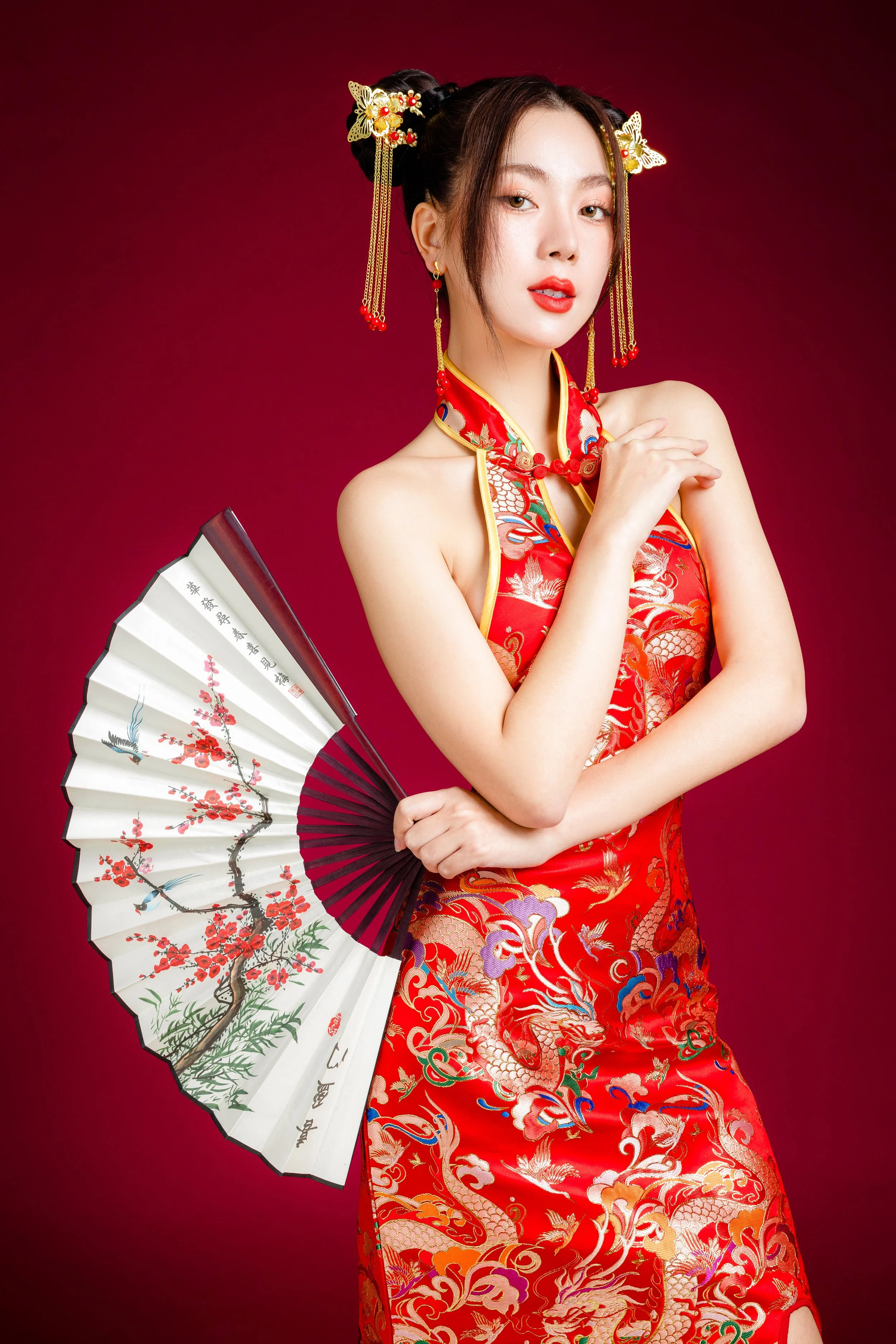 A woman dressed in a traditional Chinese red qipao with gold embroidery, holding a decorated paper fan with cherry blossom and crane designs, against a red background.