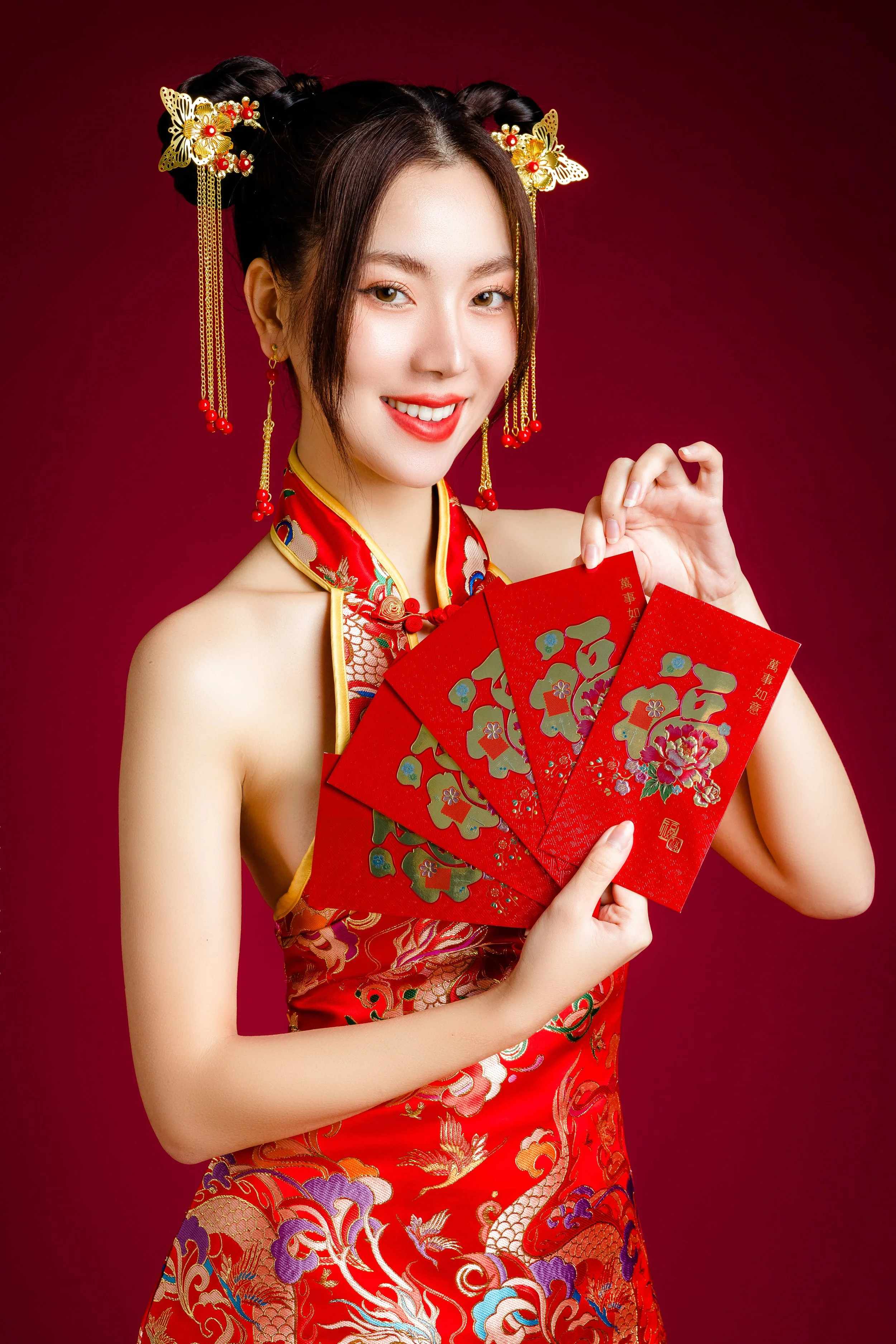 Young woman wearing traditional Chinese dress, holding red envelopes for Chinese New Year celebration, smiling against red background.