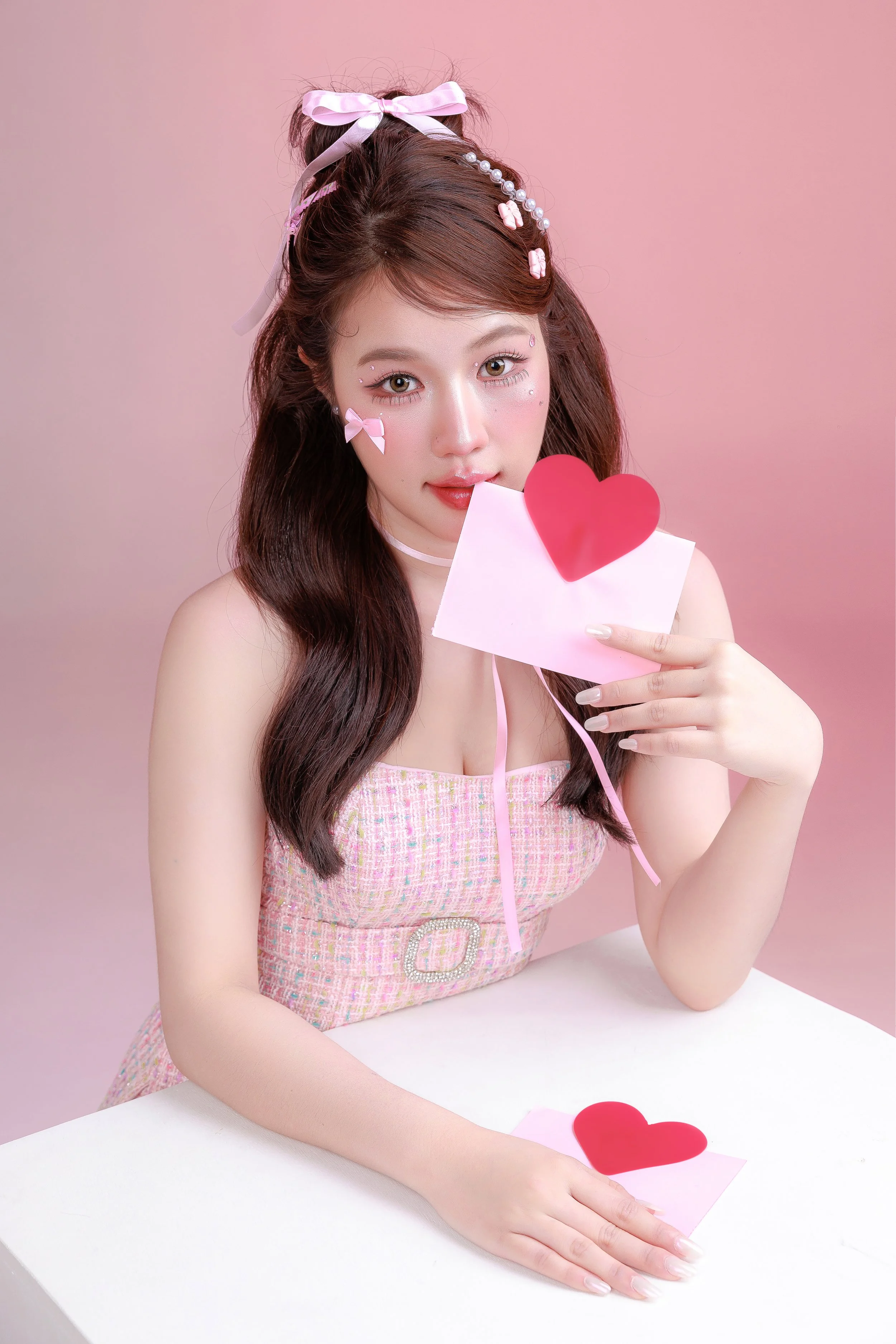 A young woman with long brown hair styled with pink bows and pearl accessories, holding a pink card with a red heart on it, sitting at a white table with another card and heart in front of her, against a pink background.