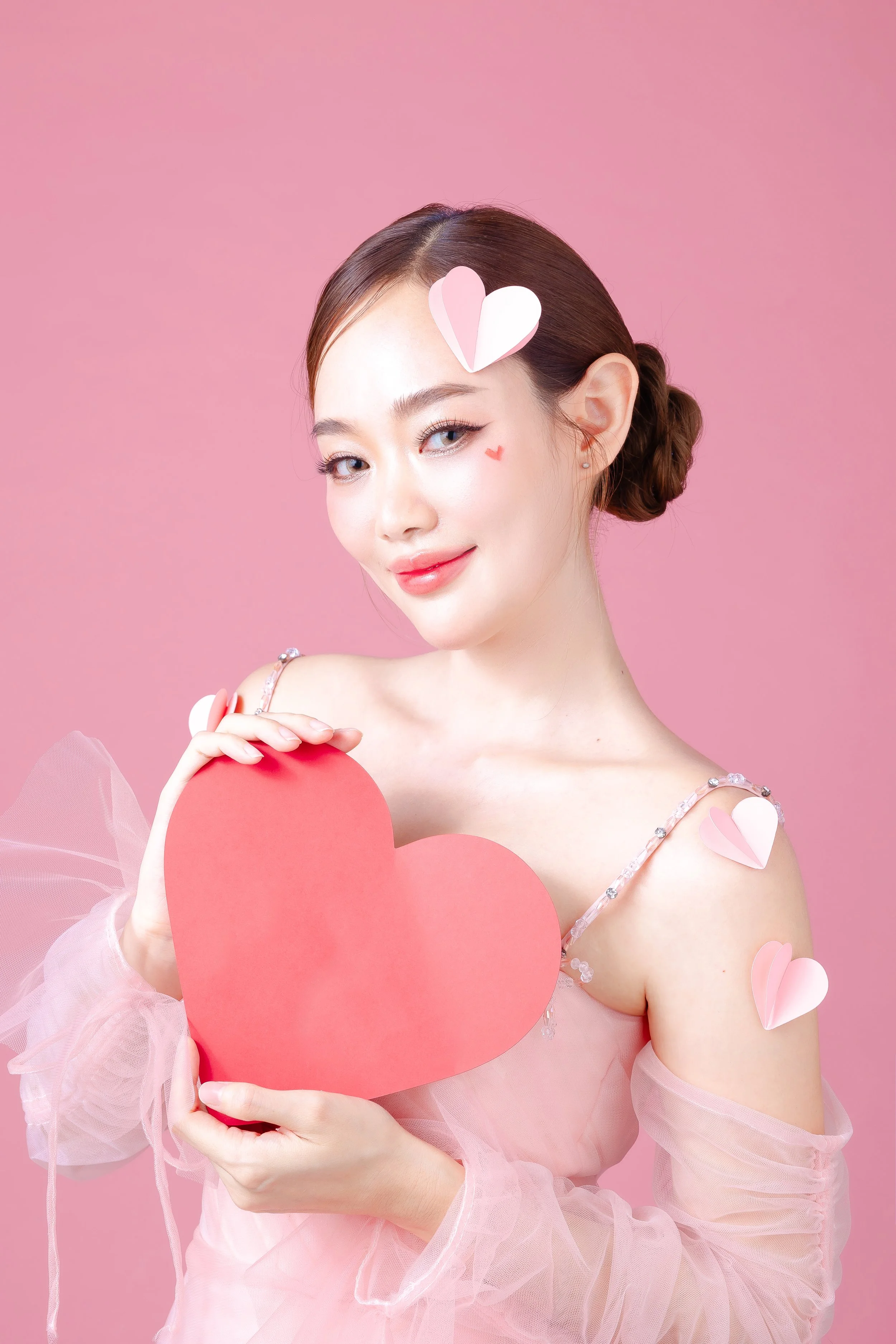 A woman with makeup and a smile, holding a large pink heart-shaped card against a pink background, decorated with paper heart stickers on her face and shoulders, wearing a pink dress with sheer sleeves.