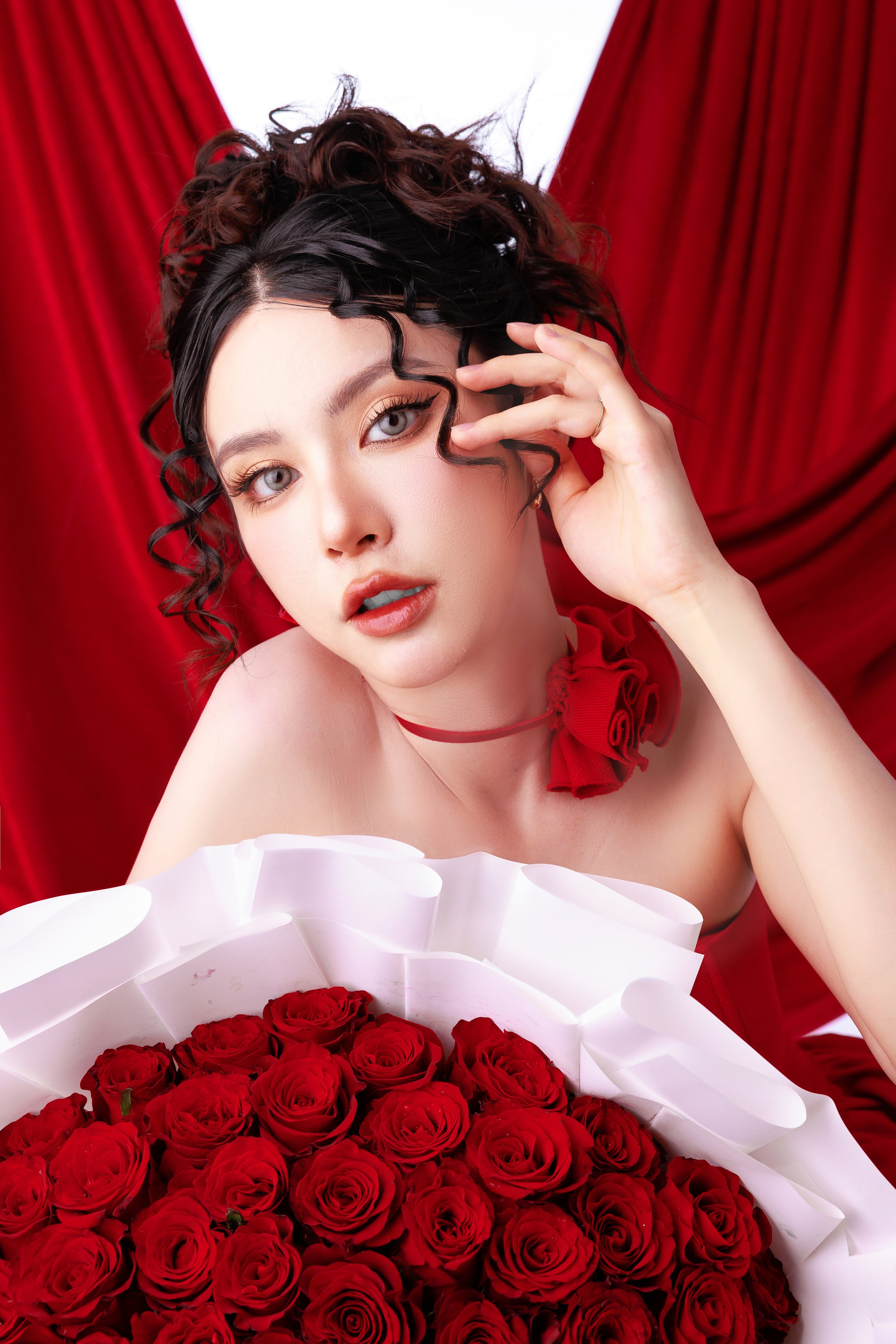 A woman with curly dark hair and blue eyes posing against red curtains, with a large bouquet of red roses in front of her.