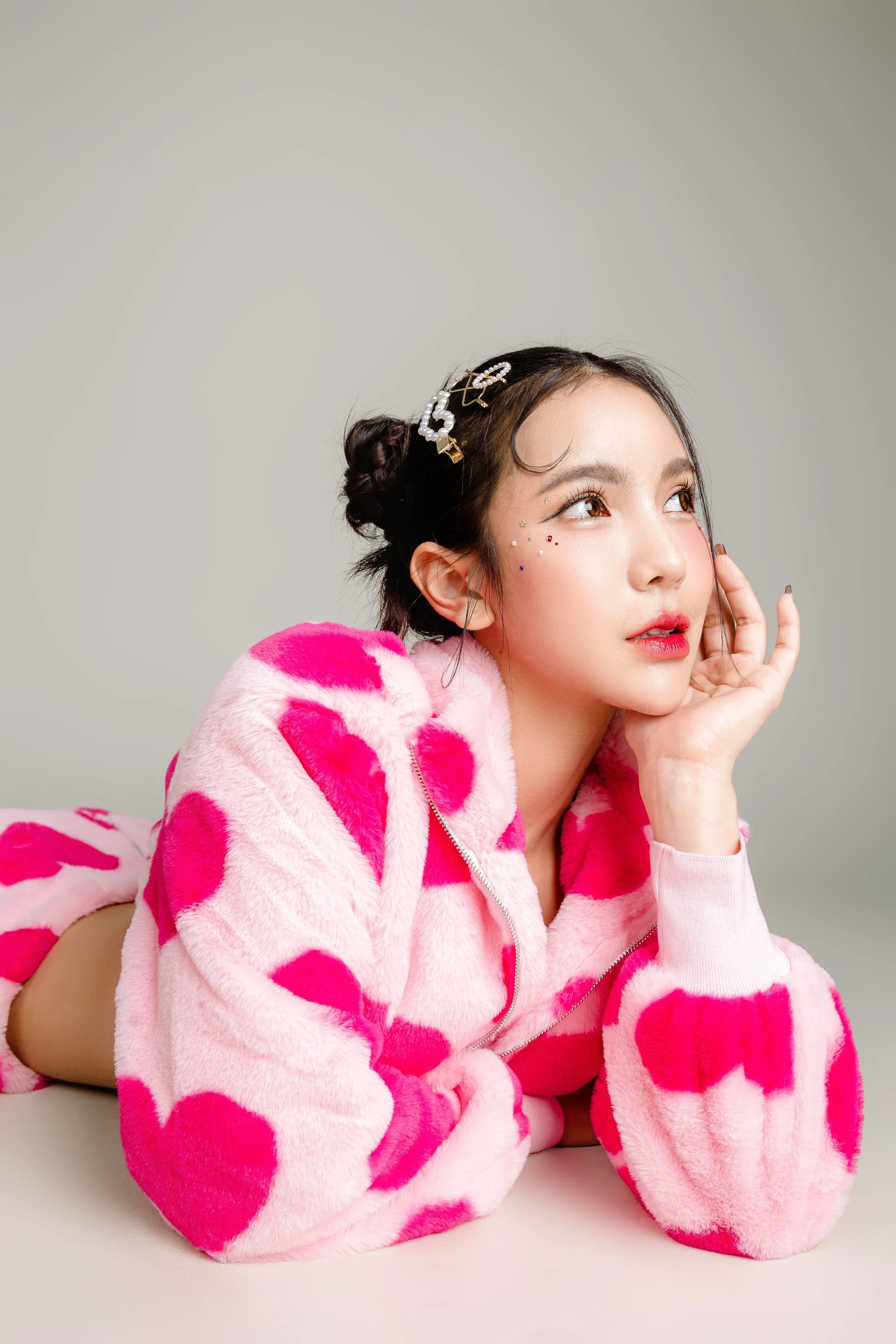 A young woman with dark hair styled in buns, wearing a pink and white furry jacket with heart patterns, lying on her stomach, resting her face on her hand, looking to the side.