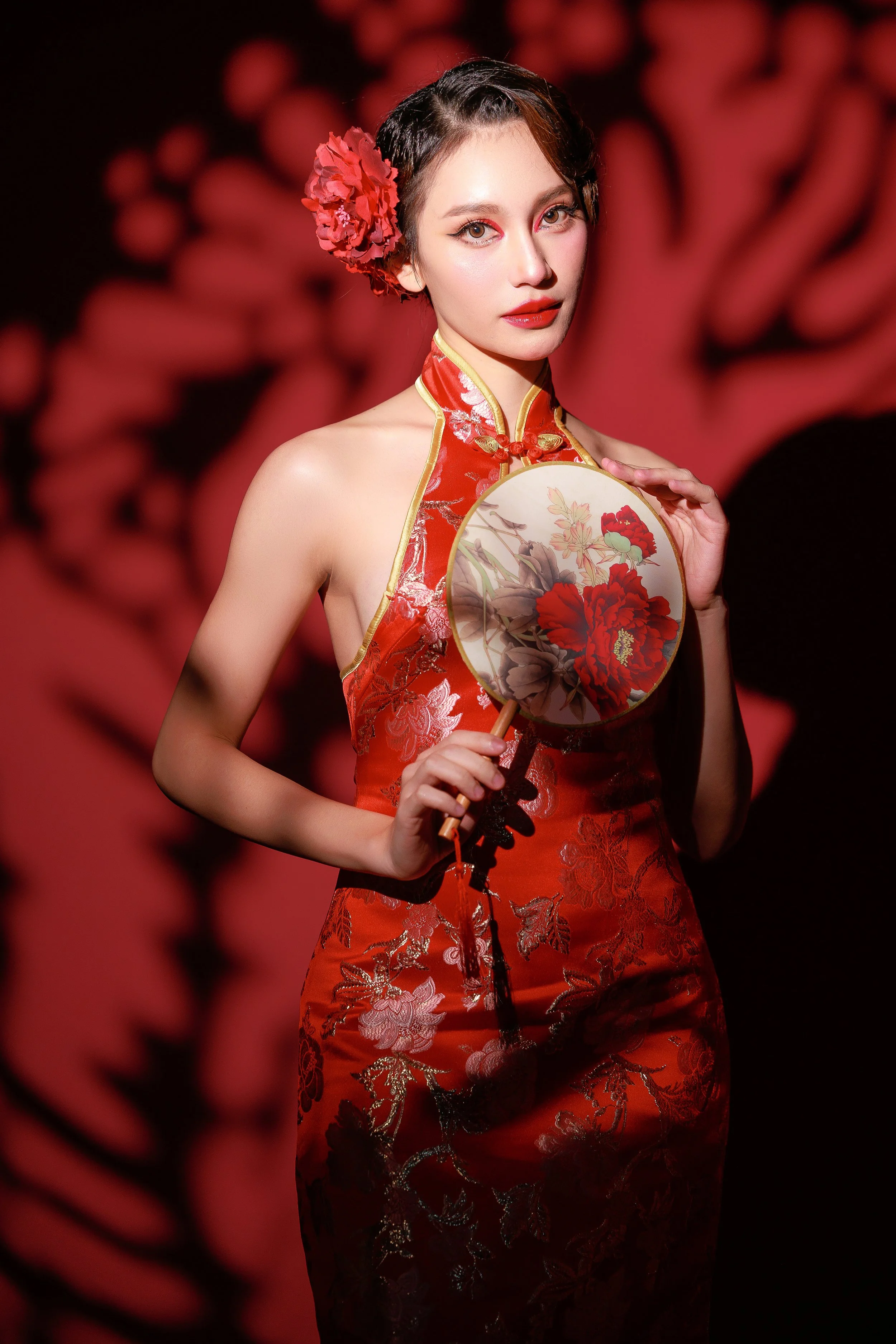A woman dressed in a red satin cheongsam with gold embroidery, holding a round fan with a peony flower design, standing against a red background with dragon motifs.