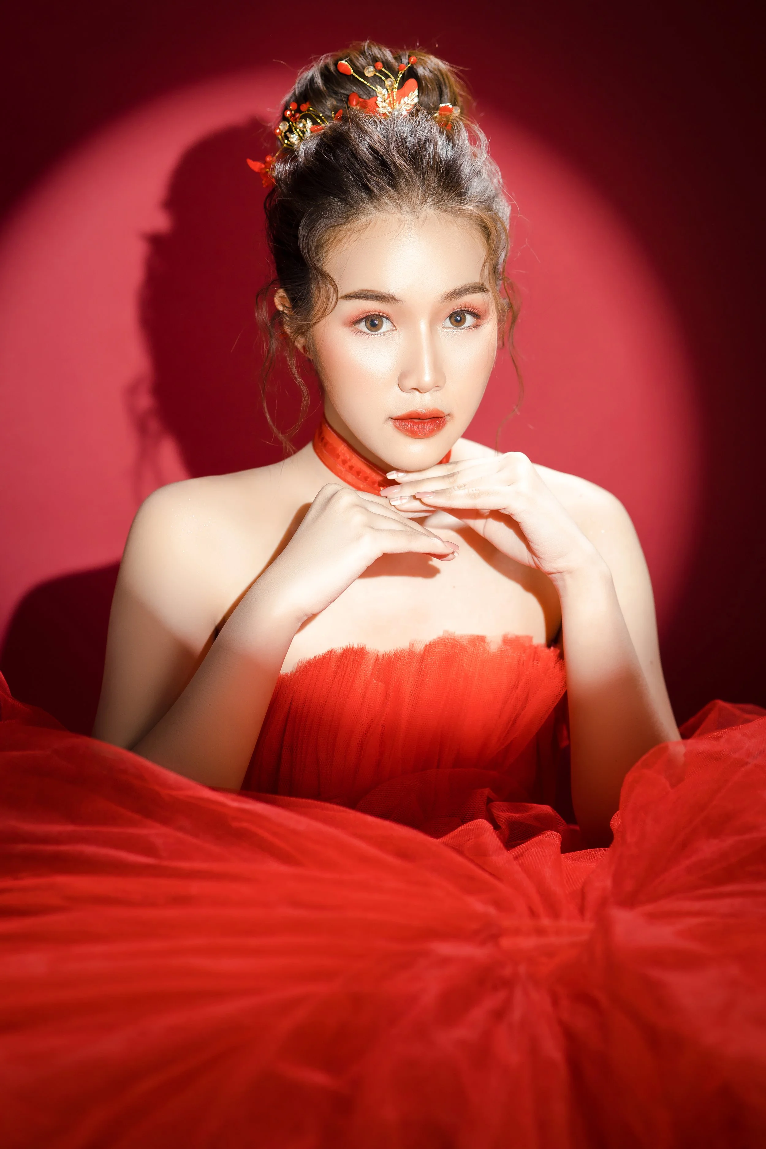 A young woman with styled hair adorned with red and gold accessories, posing against a red background. She is wearing a red strapless dress and a matching choker, with her hands gently placed near her chin, looking directly at the camera.