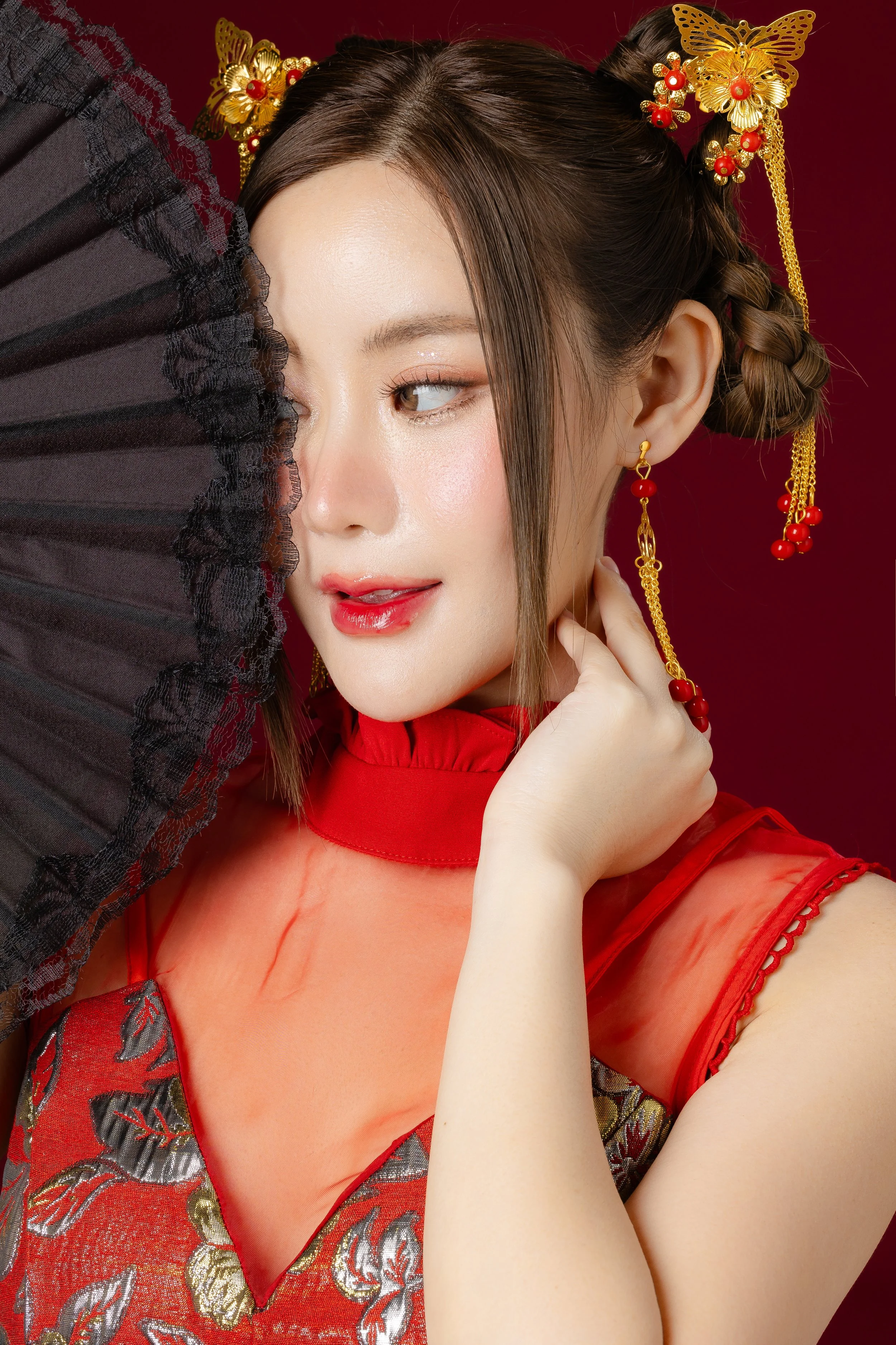 A woman with braided hair, ornate gold and red butterfly hair accessories, and red and gold earrings, holds a black lace fan partially covering her face, wearing a red embroidered dress with sheer red fabric, against a dark red background.
