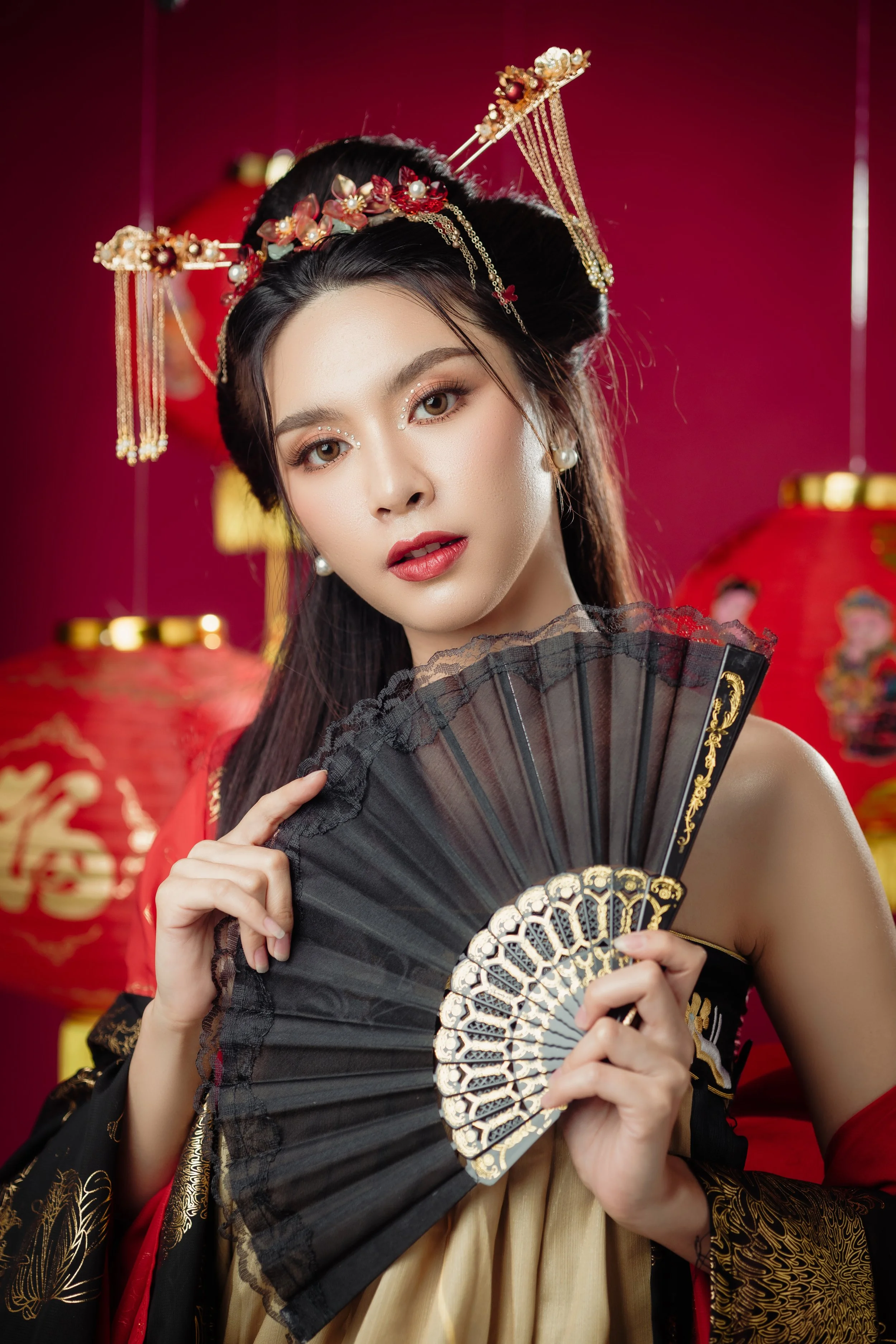 A woman in traditional Asian attire holds a black and gold folding fan in front of a red background decorated with red lanterns.
