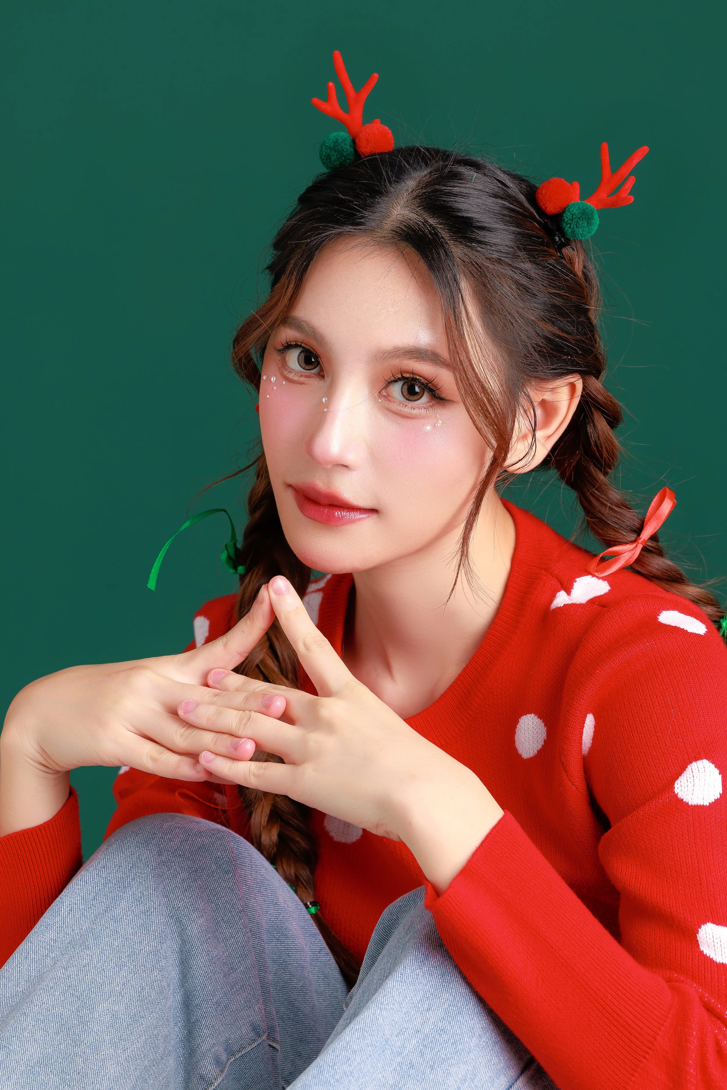 A young woman with brown hair in braids, wearing a red sweater with white polka dots, festive reindeer antlers headband, sitting against a green background.