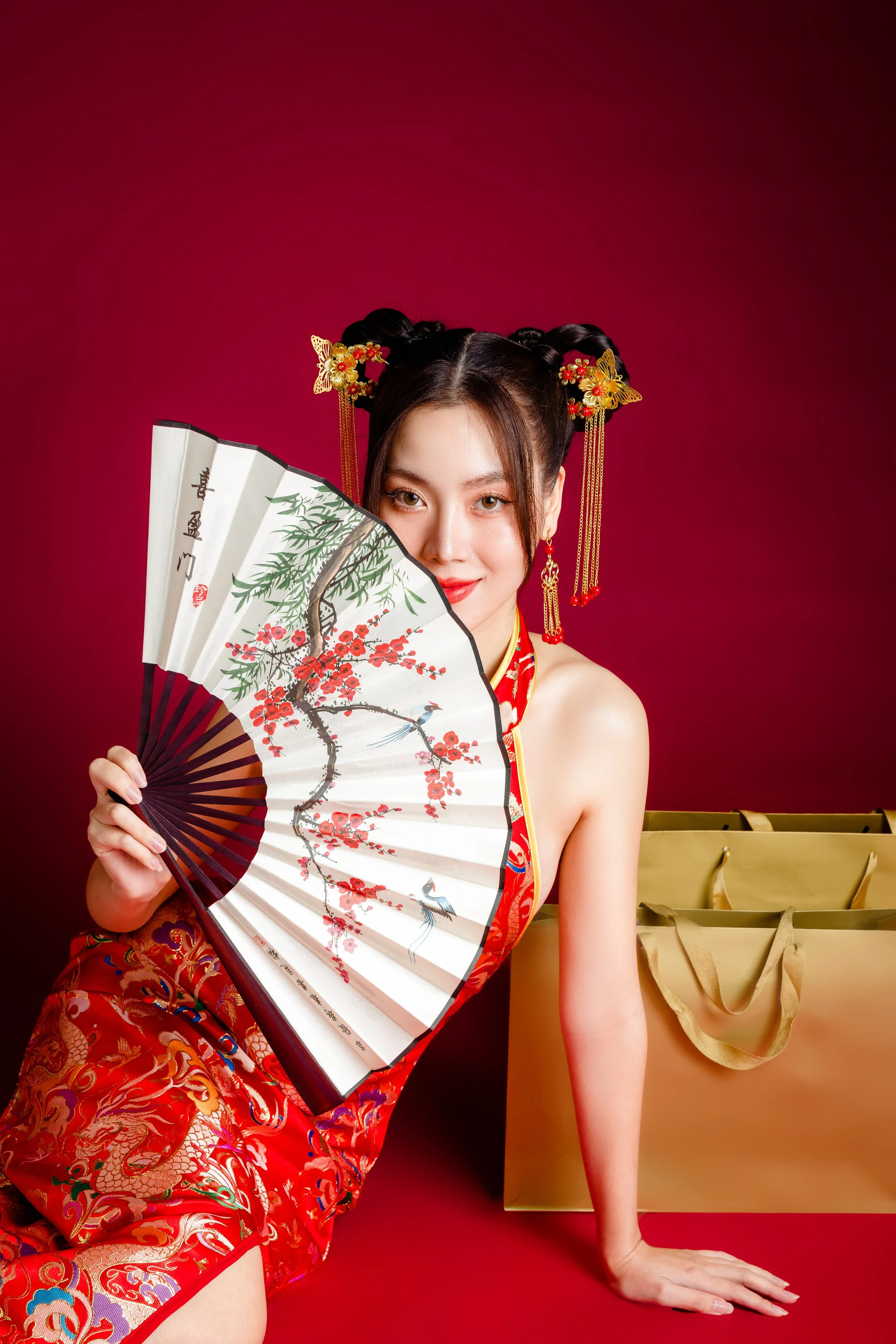 A young woman in a traditional Chinese red dress with gold and colorful embroidery, holding a decorative fan with a cherry blossom and bird design, posed against a red background with gold text and shopping bags.