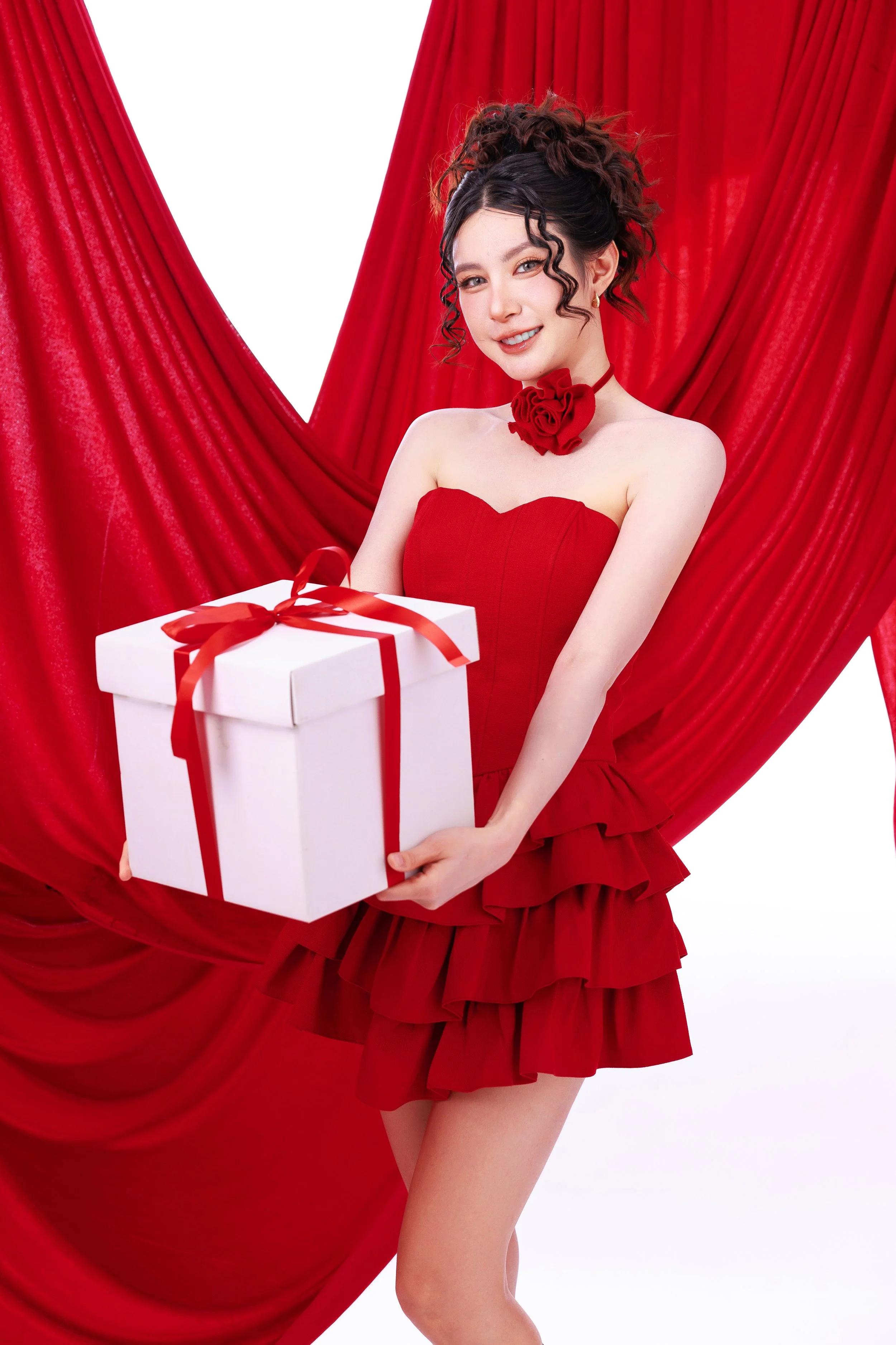 A young woman in a red dress holding a white gift box with a red ribbon, posed in front of red curtains.