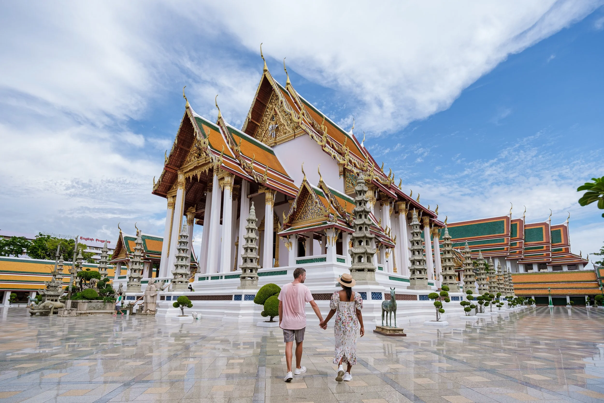 A couple in casual clothing walking hand in hand in front of a traditional Thai temple with ornate architecture, multiple tiered roofs, and decorative spires under a partly cloudy sky.