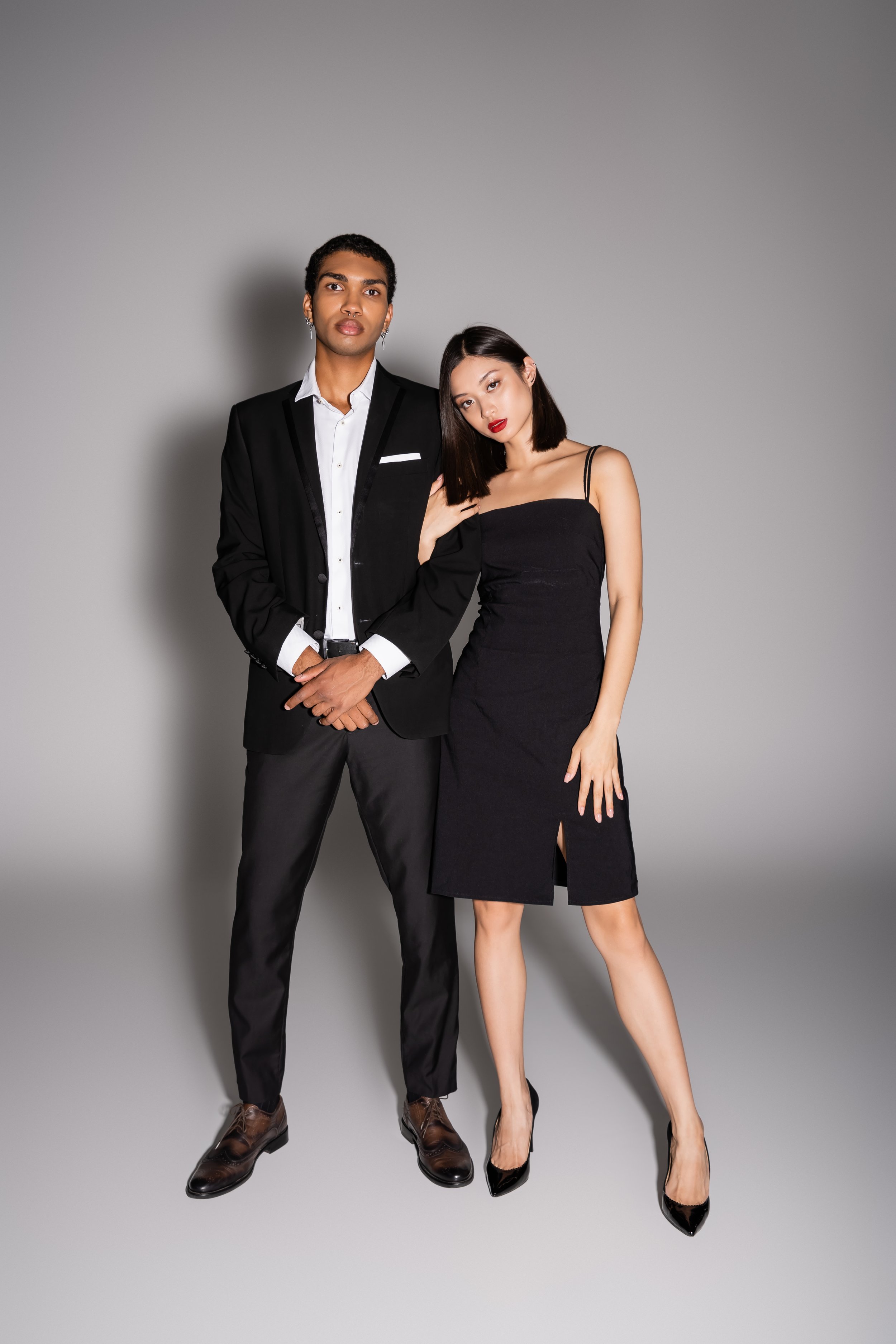 A man in a black suit and white shirt standing beside a woman in a black dress with spaghetti straps, posing together against a plain grey background.