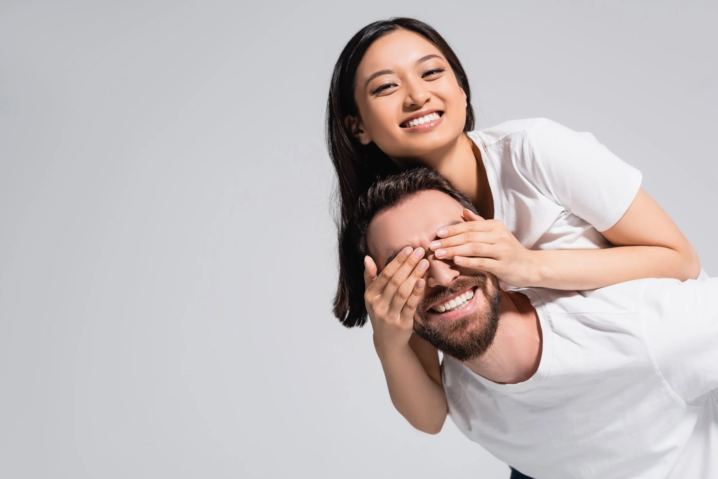 A happy couple, with the woman smiling and playfully covering the man's eyes, both wearing white t-shirts against a plain light gray background.