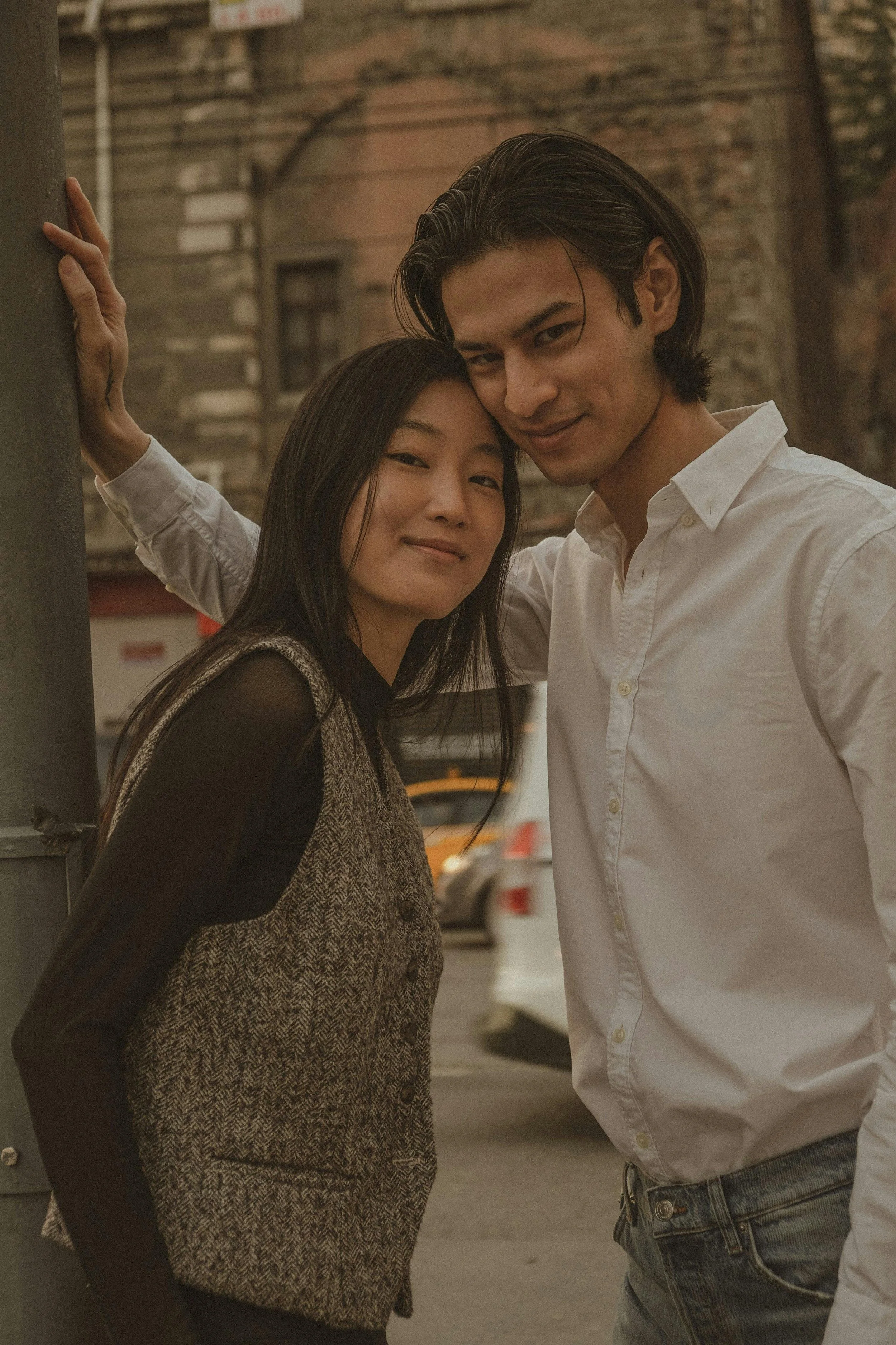 A young Asian woman and a young man with light skin posing closely together on a city street during dusk, with their heads touching and smiling softly, with cars and old brick buildings in the background.