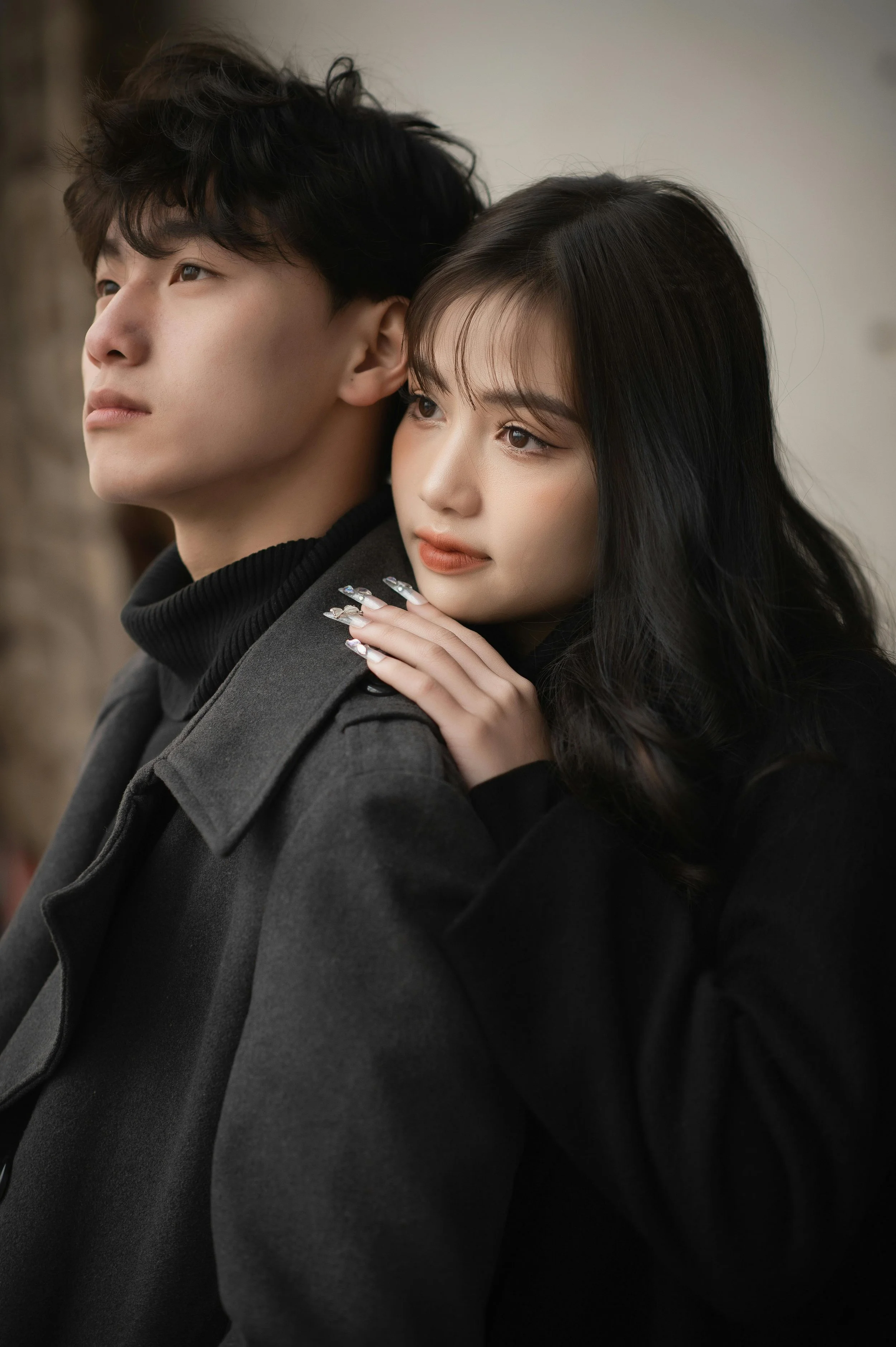 A young man and woman in black clothing with the woman resting her head on the man's shoulder and her hand on his shoulder, both gazing into the distance.