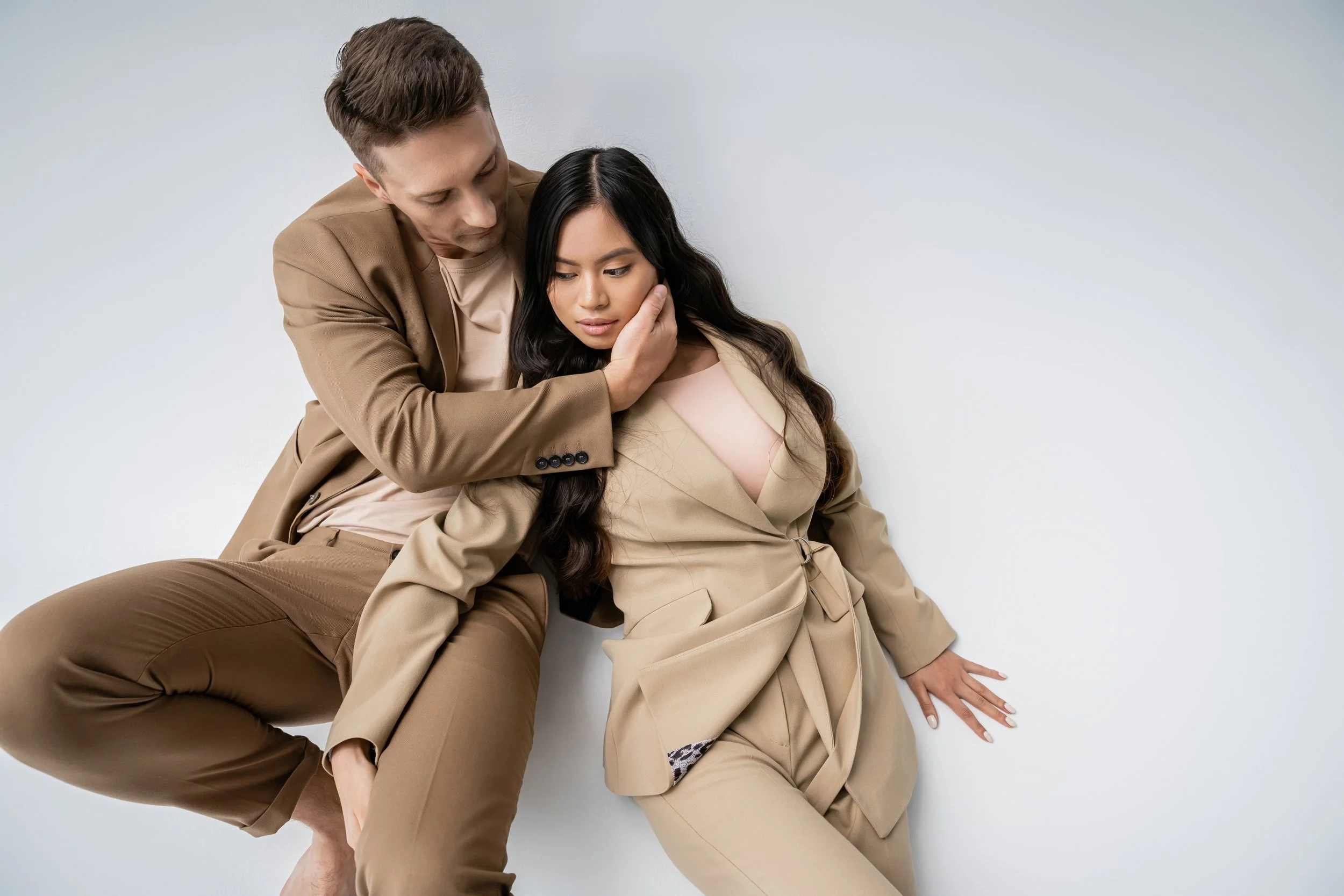 A man and woman in beige suits posed against a plain white background, with the man gently holding the woman's face as she leans back.