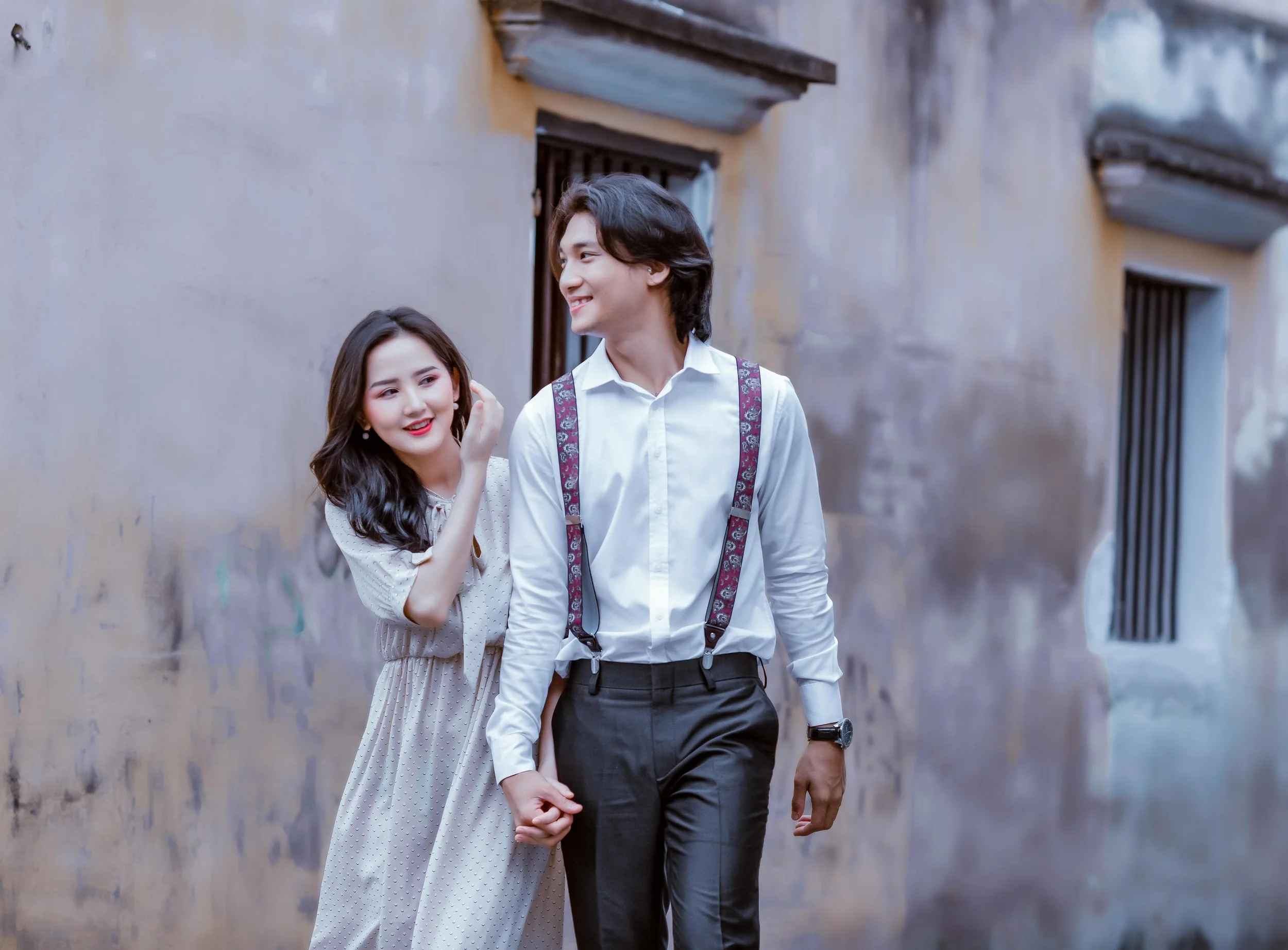 A young couple walking hand in hand and smiling, holding hands, in an alley with a rustic wall in the background. The woman has long dark hair and is wearing a light-colored dress, while the man has dark hair and is dressed in a white shirt with susp