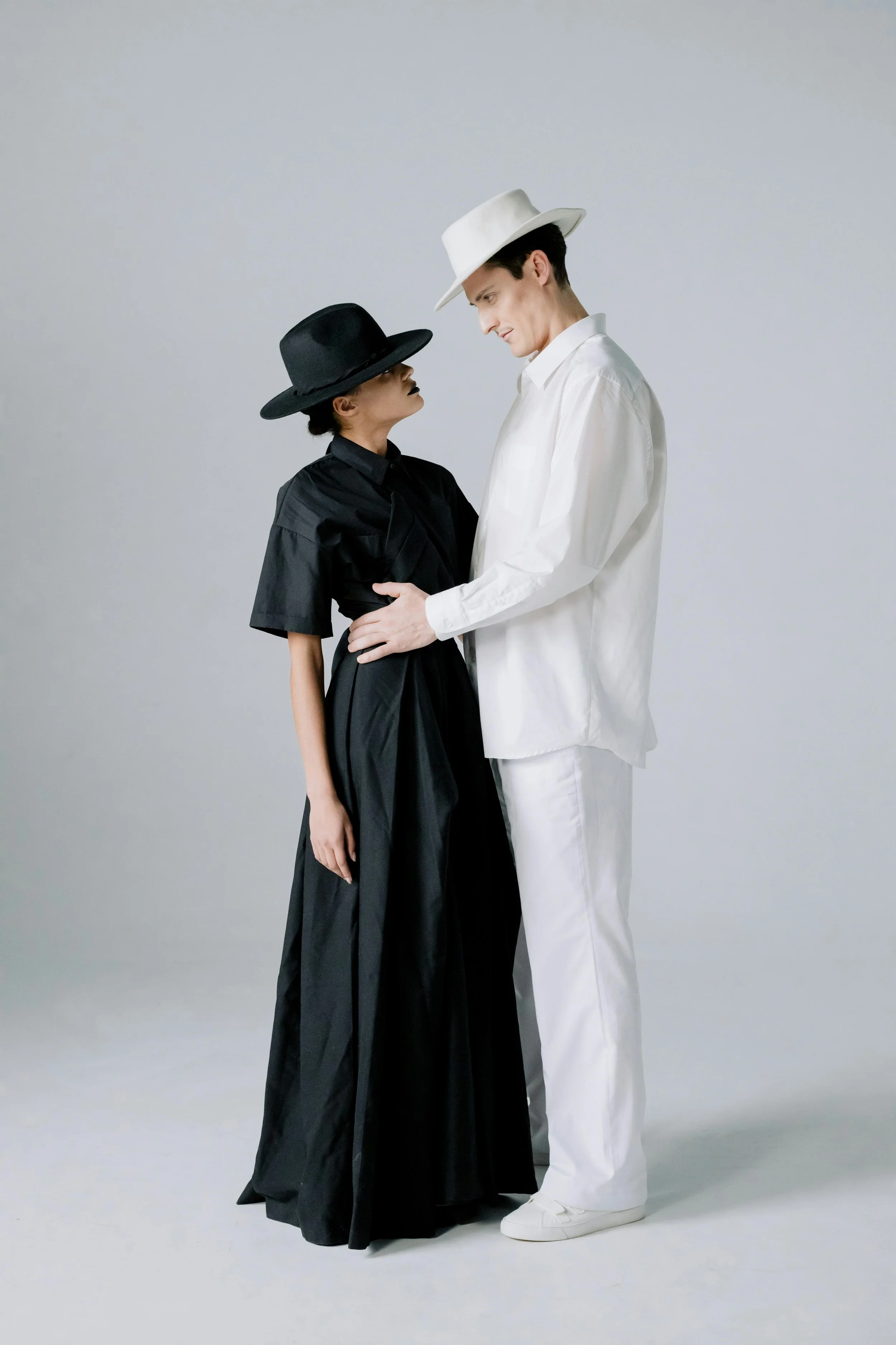 A man and woman facing each other, dressed in black and white clothing with hats, standing close in a minimalist setting.