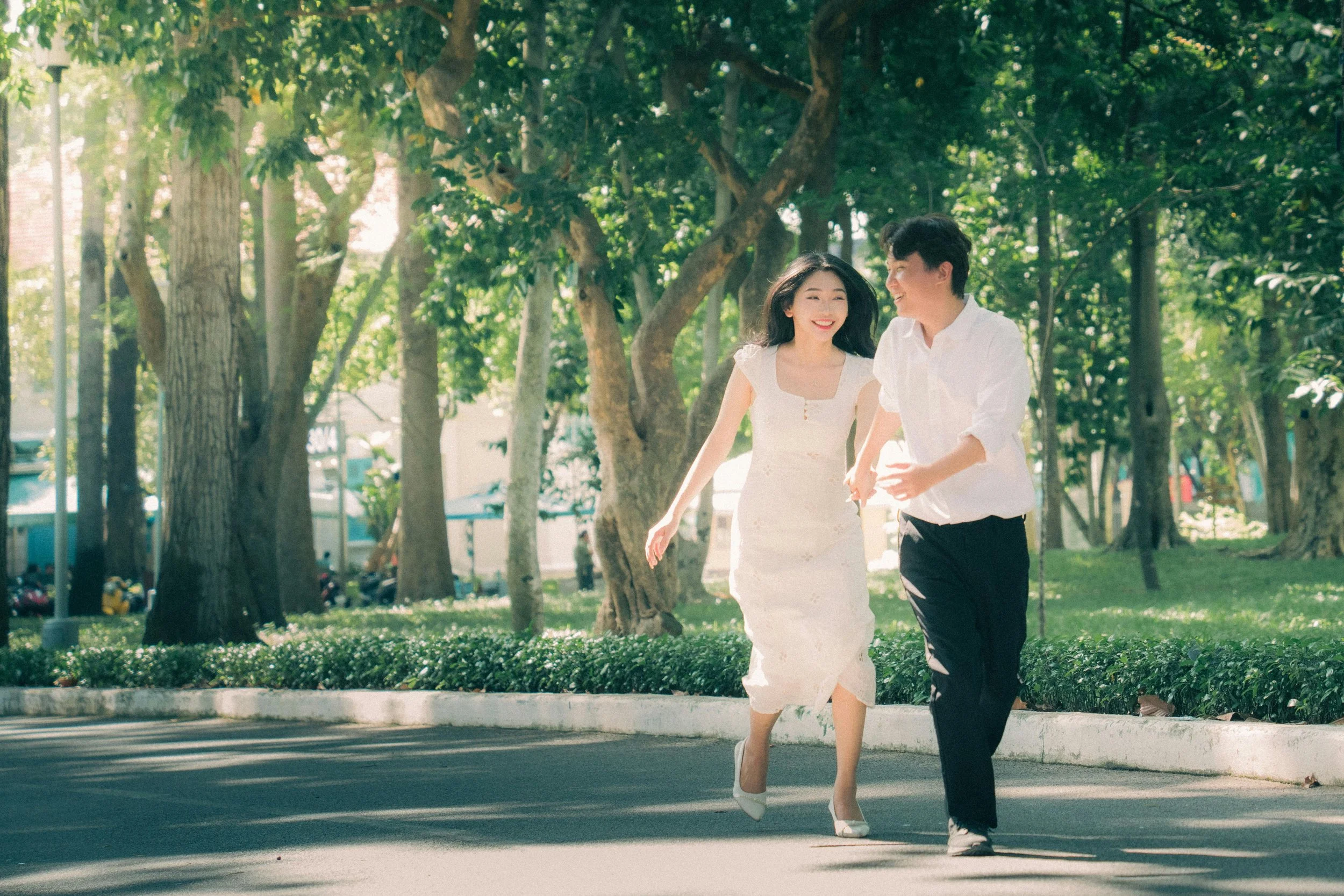 A couple running on a tree-lined sidewalk in a park, smiling and holding hands, with sunlight filtering through the leaves.