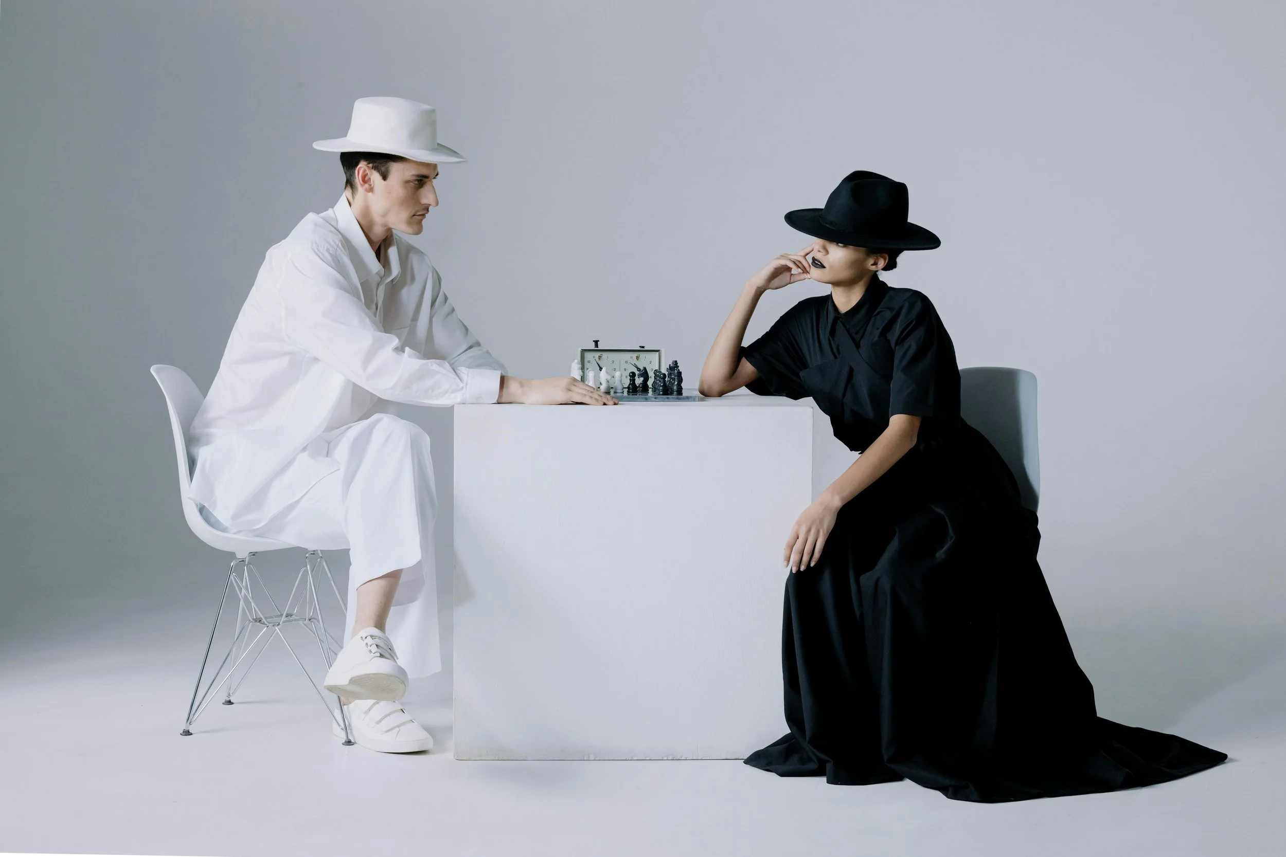 A man and woman sit across from each other at a white table, playing chess. The man is dressed in all white, wearing a hat, and the woman is dressed in all black, also wearing a hat. The woman is resting her chin on her hand, contemplating her move.