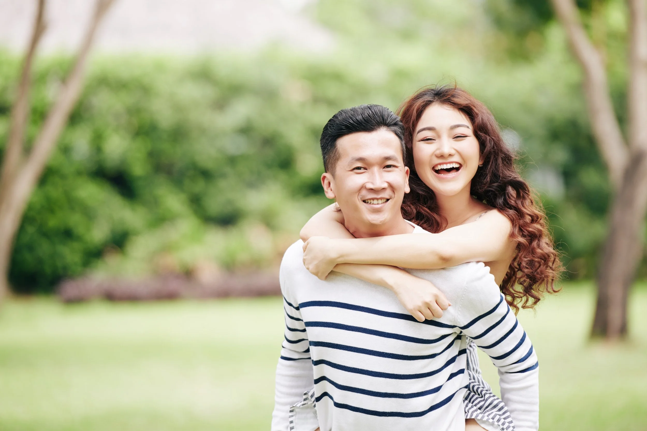 A happy couple is outdoors in a park, with the woman giving the man a piggyback ride. Both are smiling and enjoying the moment, surrounded by green trees and grass.