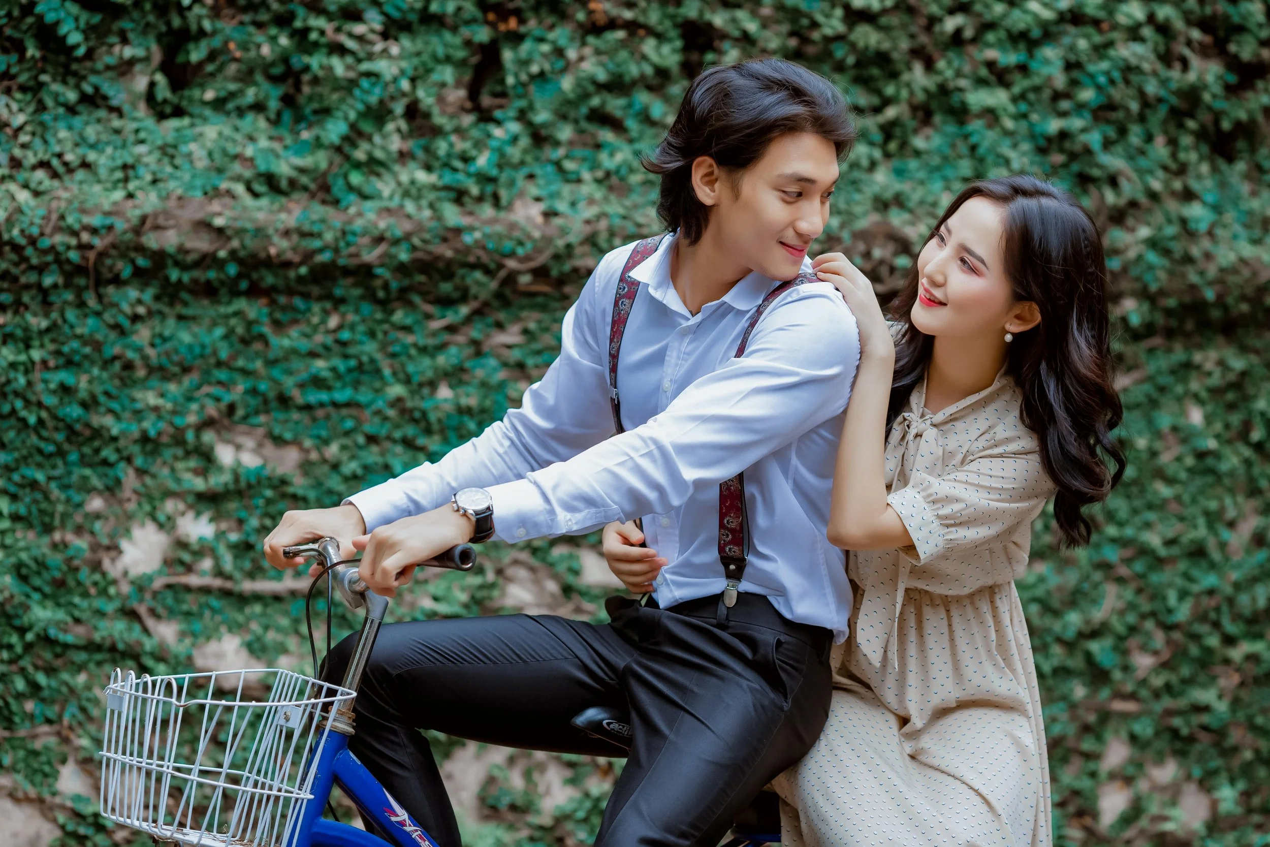 A young couple enjoying a moment outdoors, with the man riding a bicycle and the woman sitting behind him, smiling and touching his shoulder, against a background of green leafy plants.