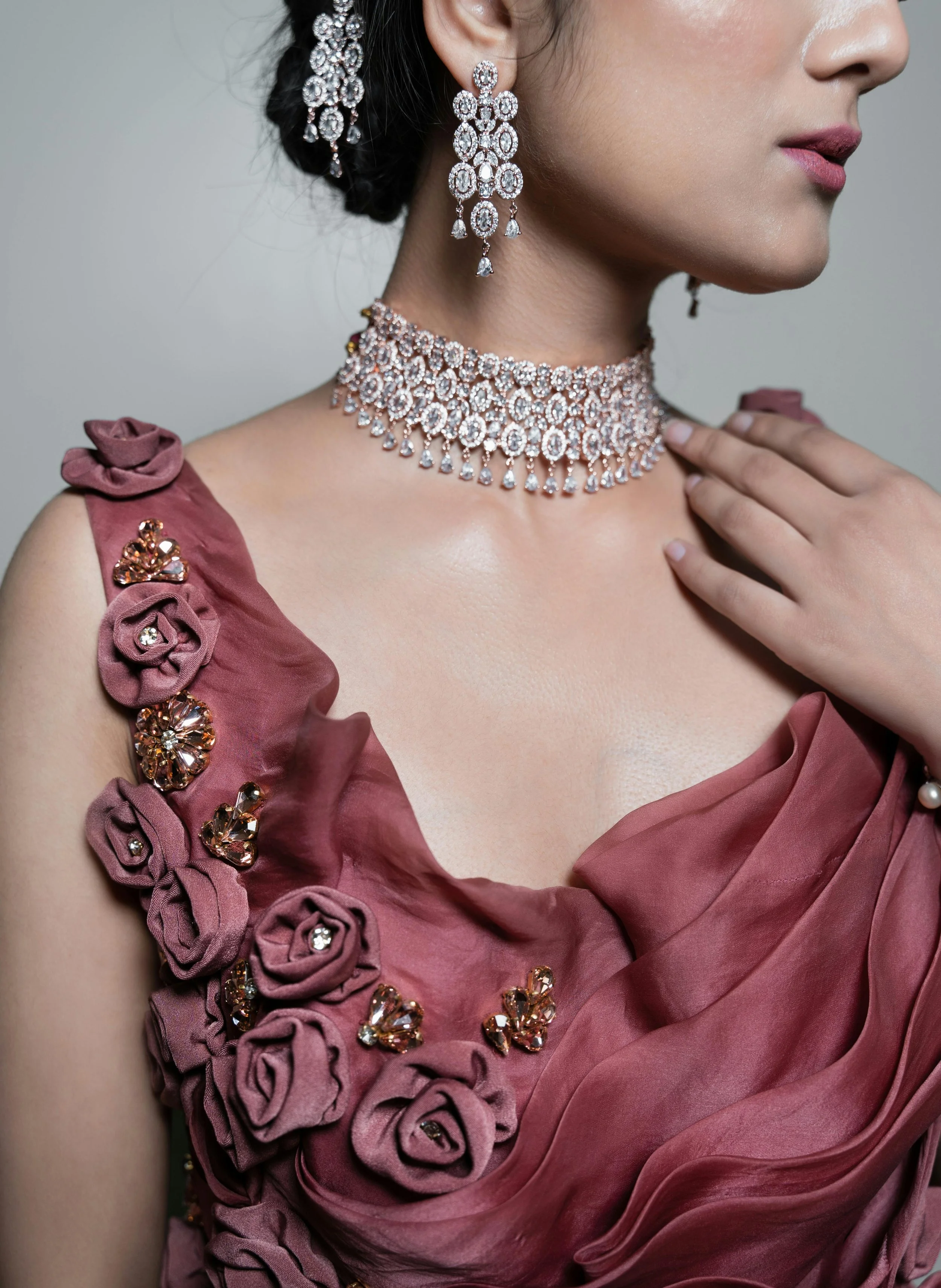 Close-up of a woman wearing elegant jewelry and a dress. She has diamond earrings, a diamond choker, and a pearl bracelet. Her dress is pink with fabric flowers and embellishments on one shoulder.