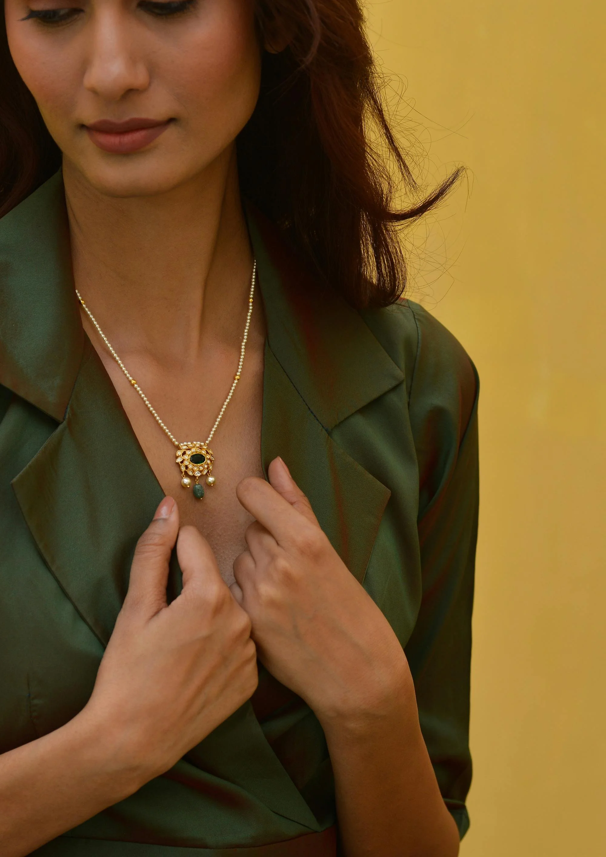 Close-up of a woman wearing a green satin blouse, holding a necklace with a pendant featuring a black gemstone and decorative elements, against a yellow background.