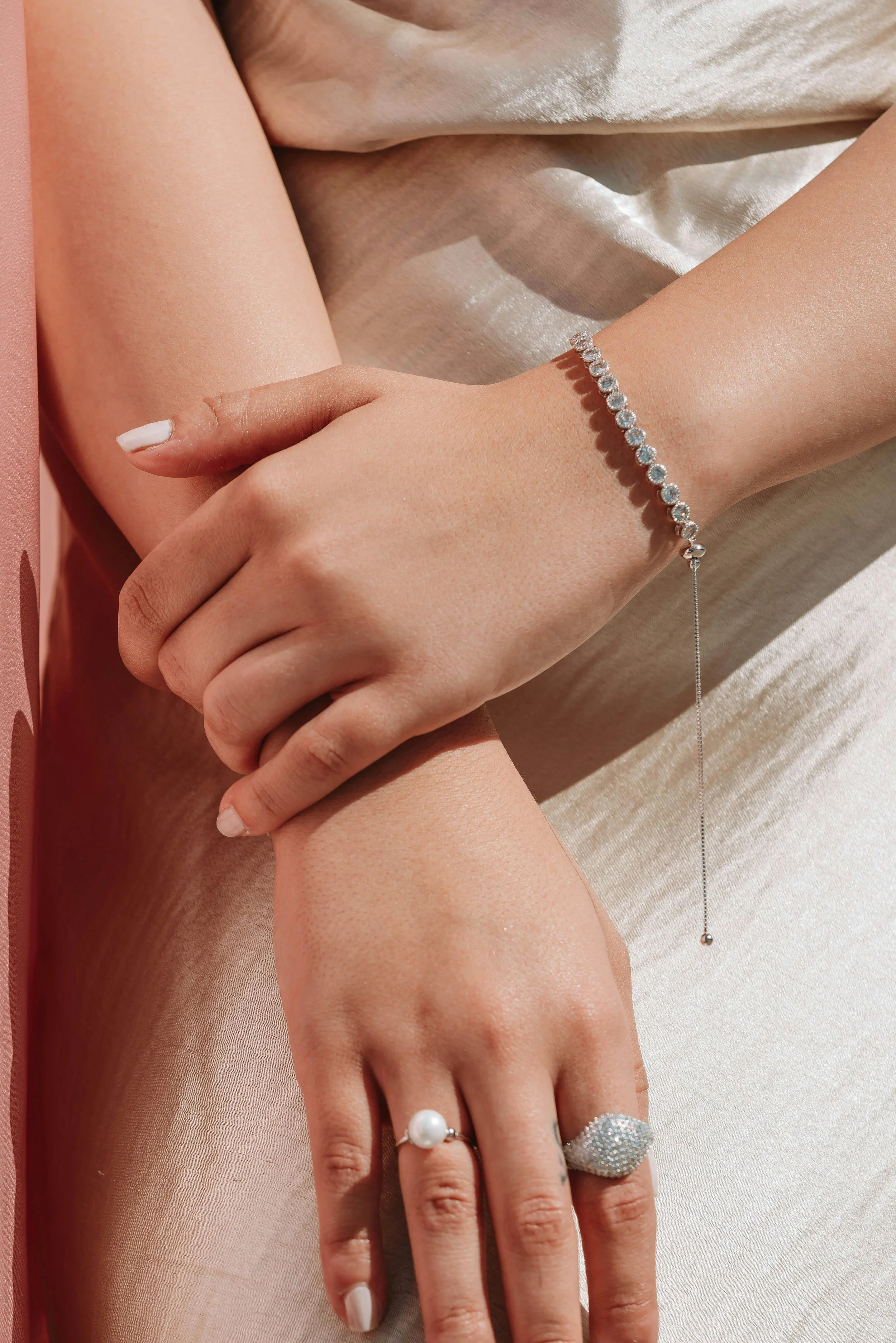 Close-up of a woman's hands and wrists showing jewelry, including a pearl ring, a large diamond-like ring, a diamond bracelet, and a delicate chain bracelet with a small ball at the end. The woman is resting her hands on her knee, which is draped in 