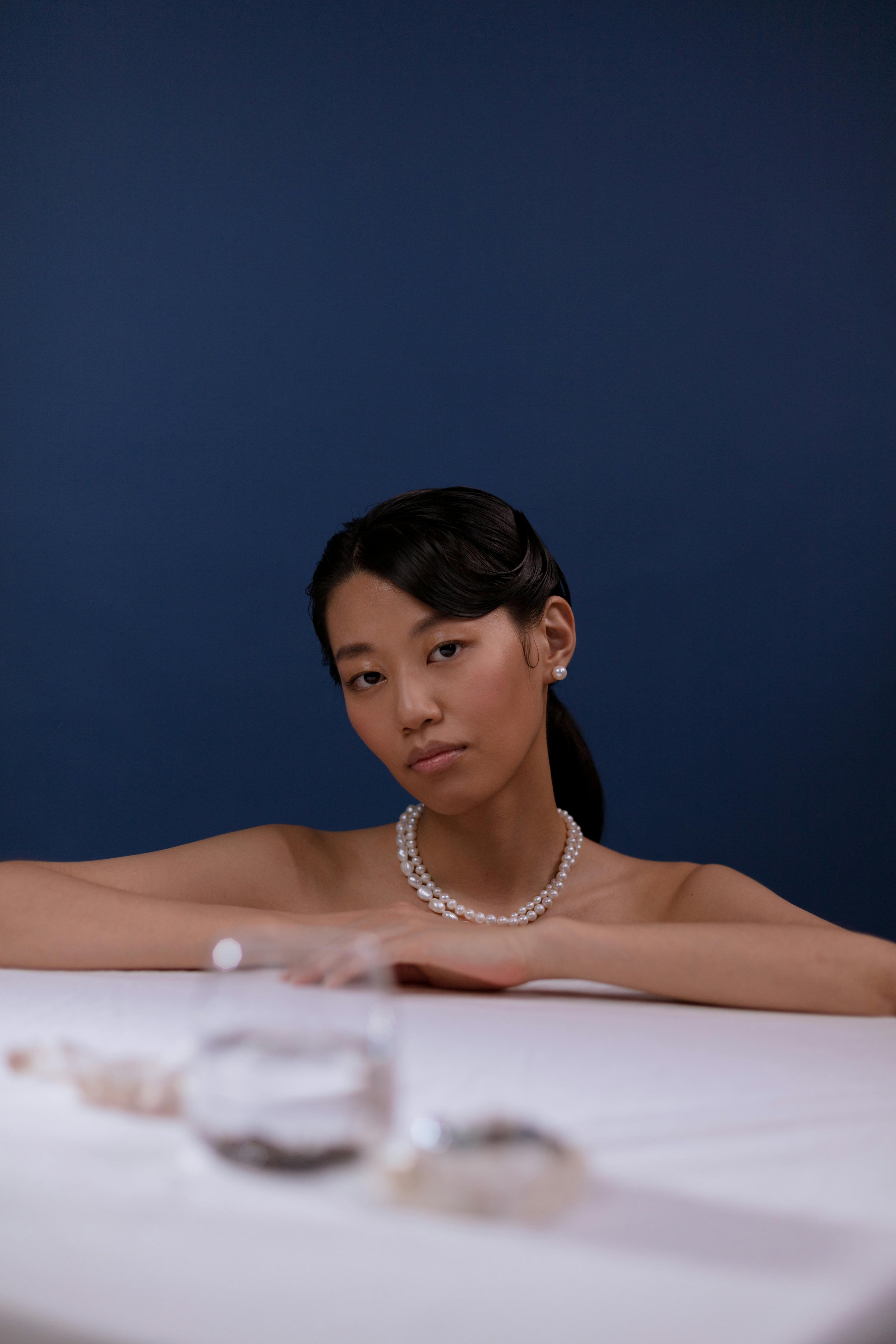 A woman with dark hair wearing pearl jewelry, resting her arms on a table, with a blue background.
