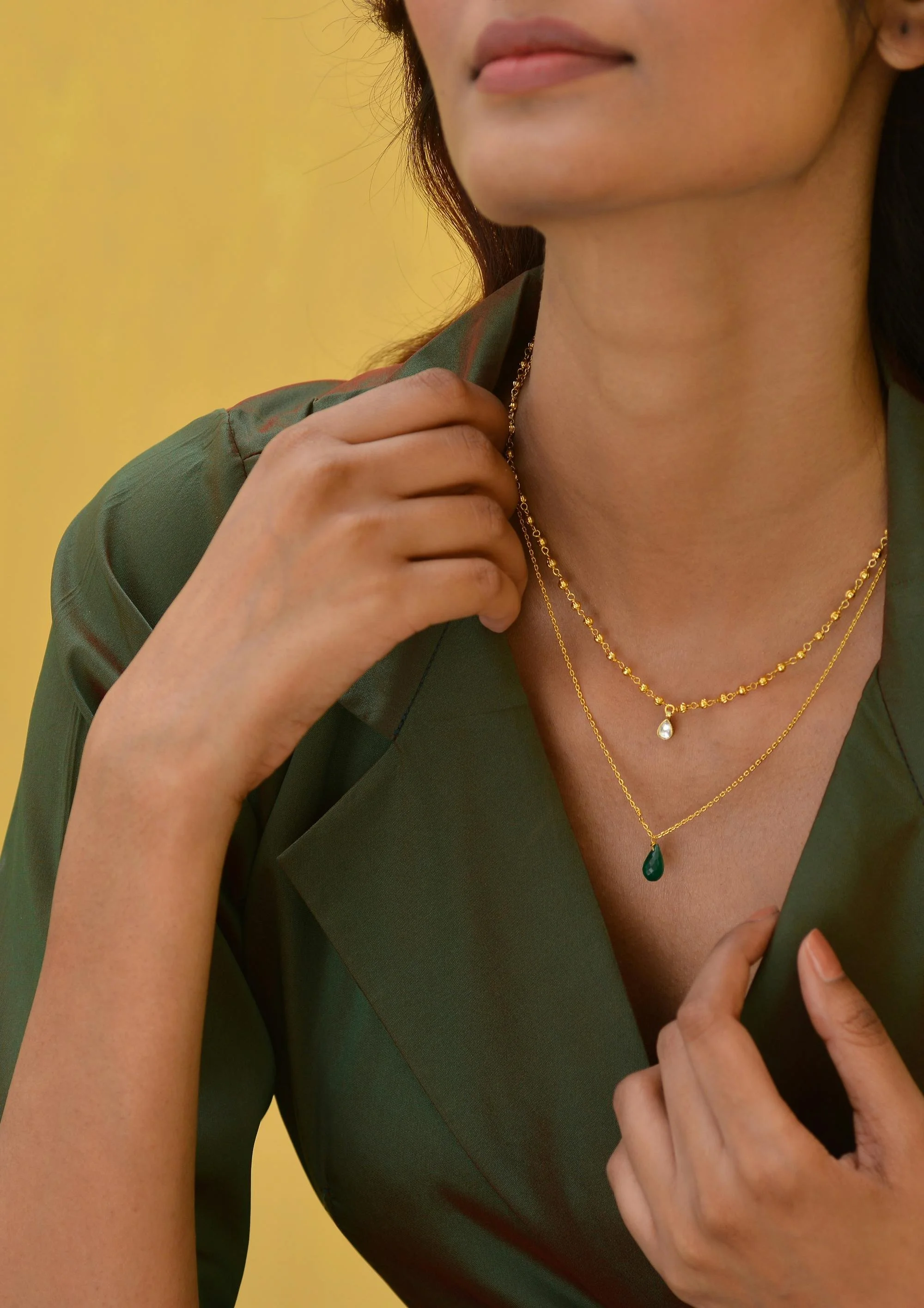 Close-up image of a woman wearing layered gold necklaces with teardrop pendants, dressed in a green blazer, with a yellow background.