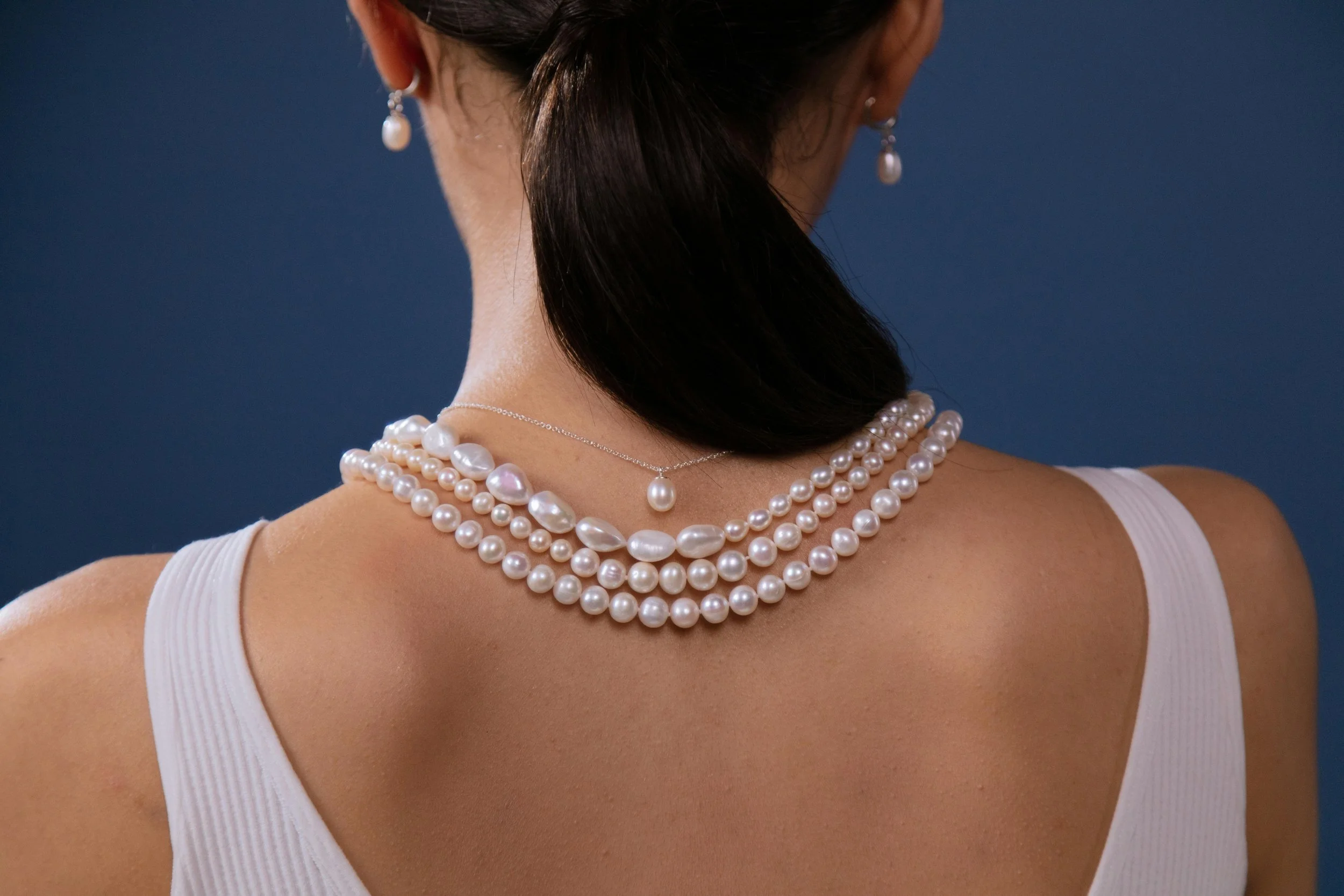 Back of a woman's neck and shoulders showing she is wearing multiple strands of pearl necklaces and pearl earrings, with her dark hair styled back and a white sleeveless top against a dark blue background.