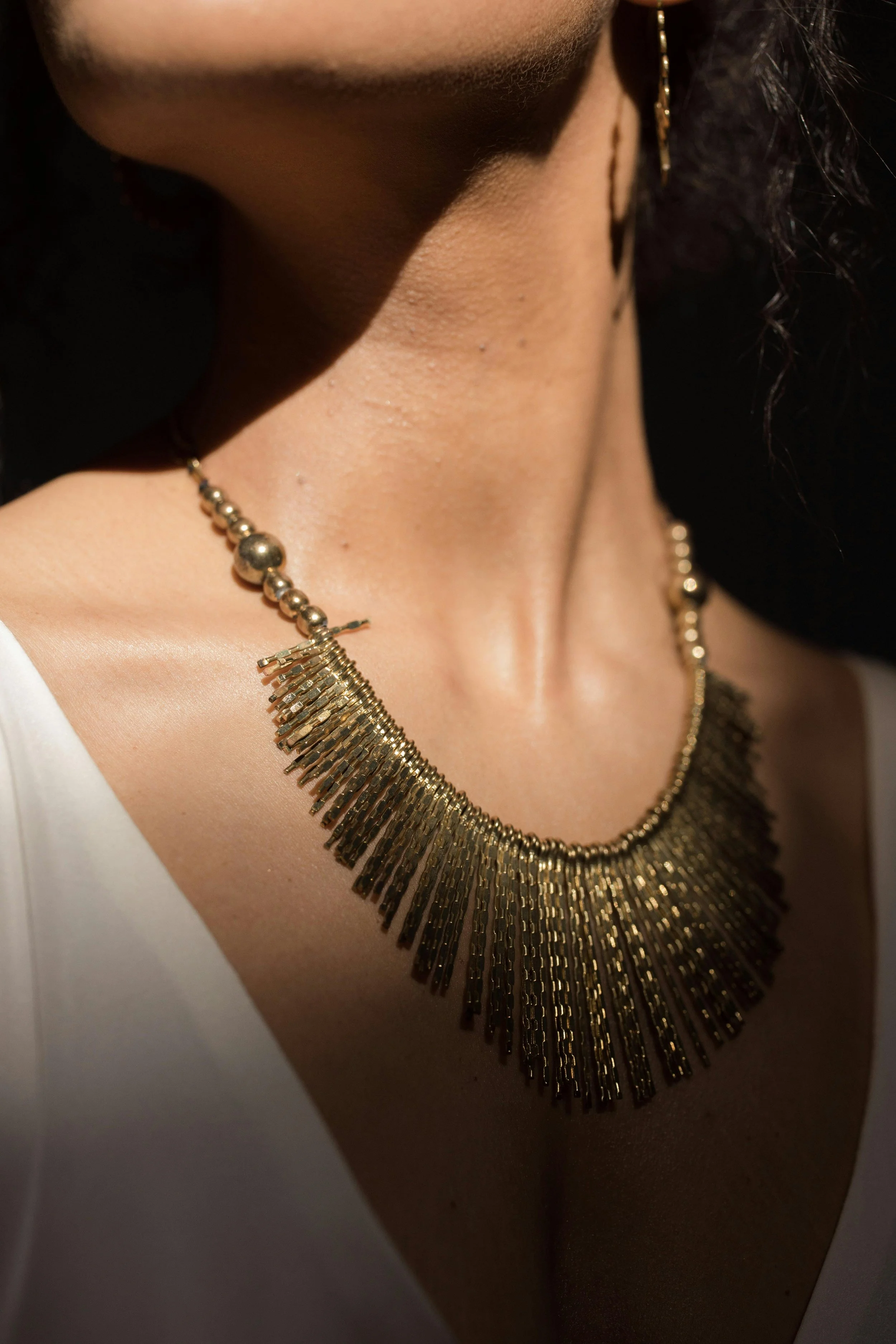 Close-up of a woman's neck and shoulder adorned with a gold statement necklace with fringe details, a small part of her white top, and an earring visible on the right side.