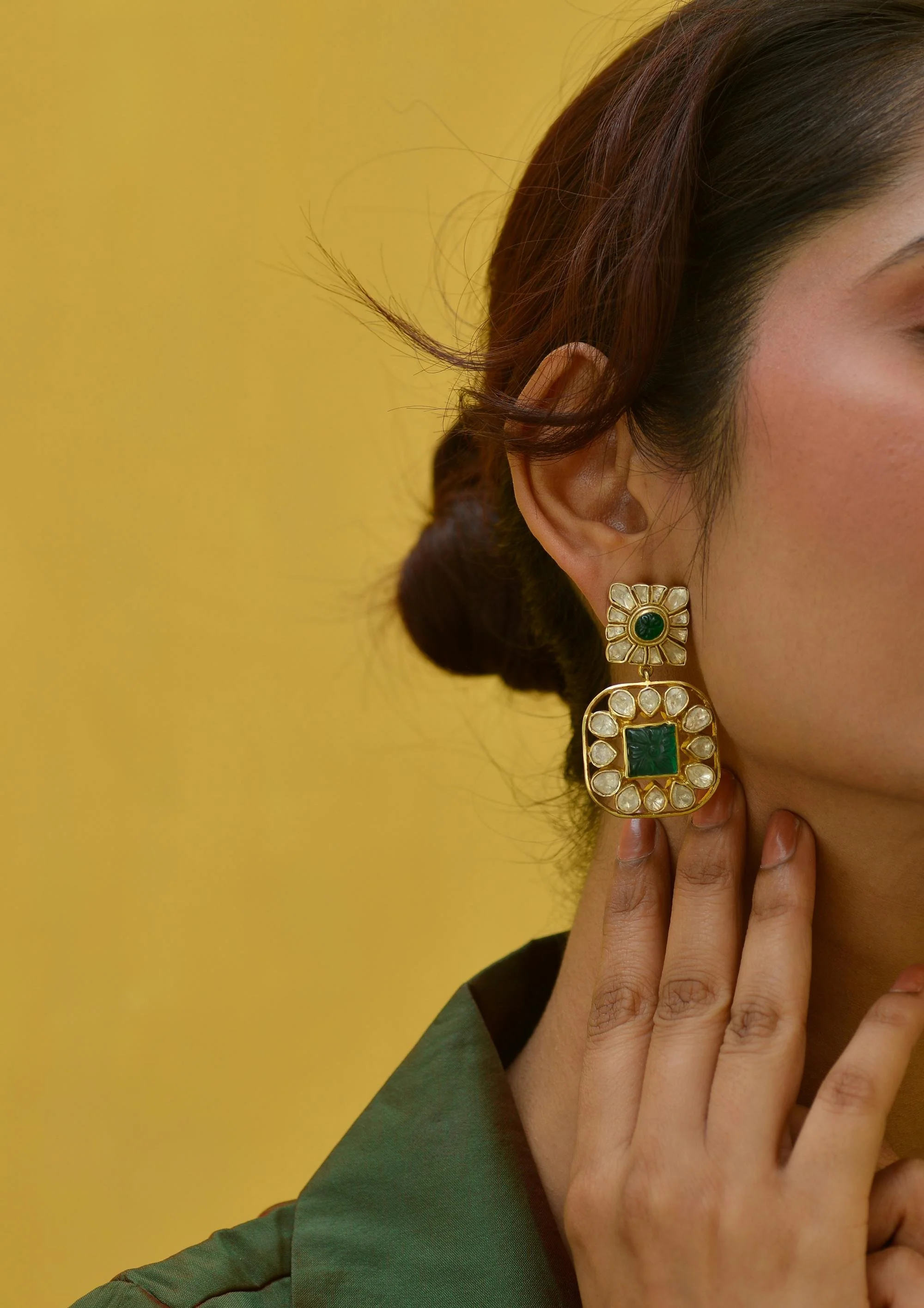 Close-up of a woman's face showing her eye, hairstyle, and an ornate large gold earring with green and white stones, set against a yellow background.
