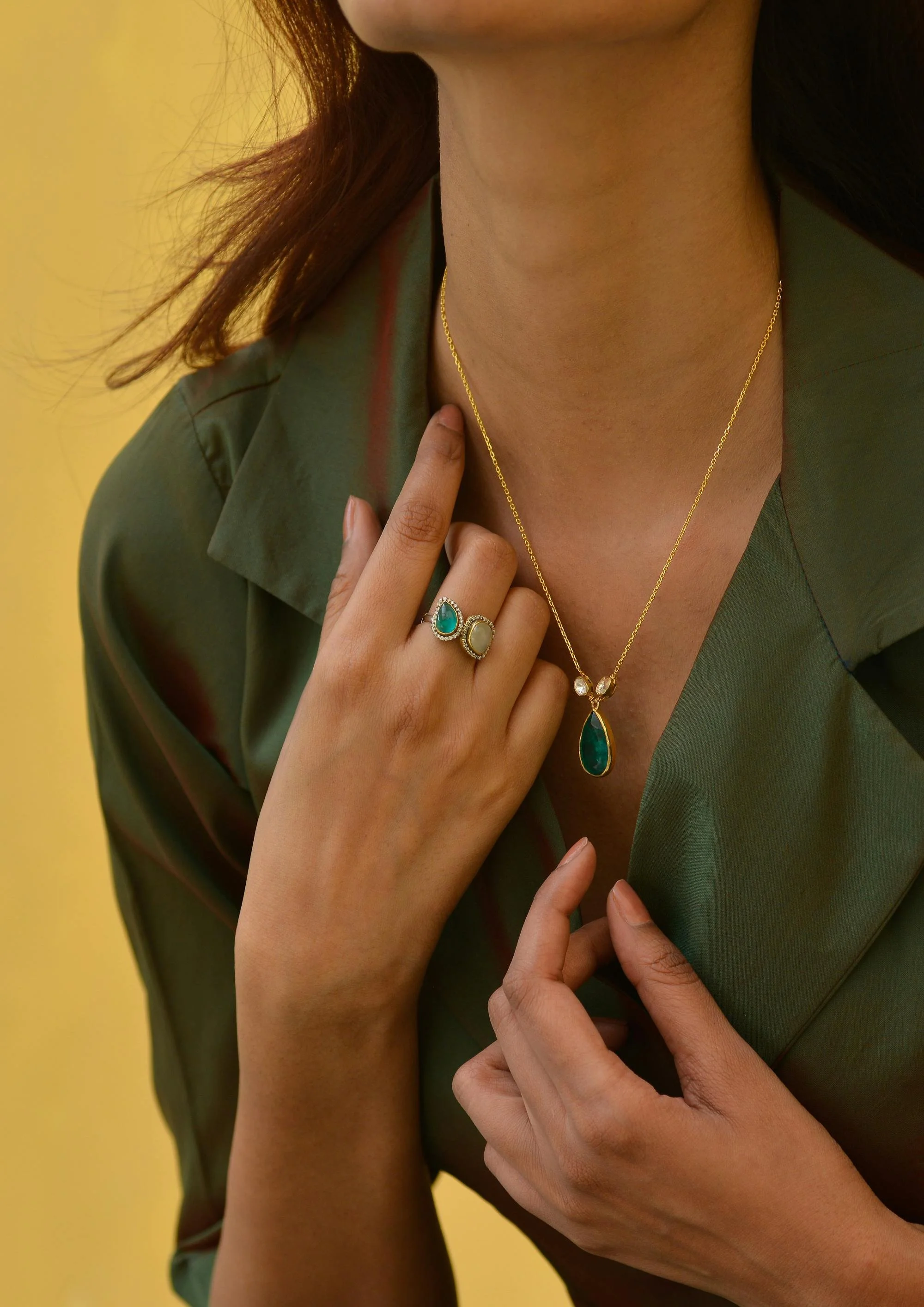 Close-up of a woman wearing gold jewelry, including two rings and a necklace, posed against a yellow background and dressed in a green shirt.
