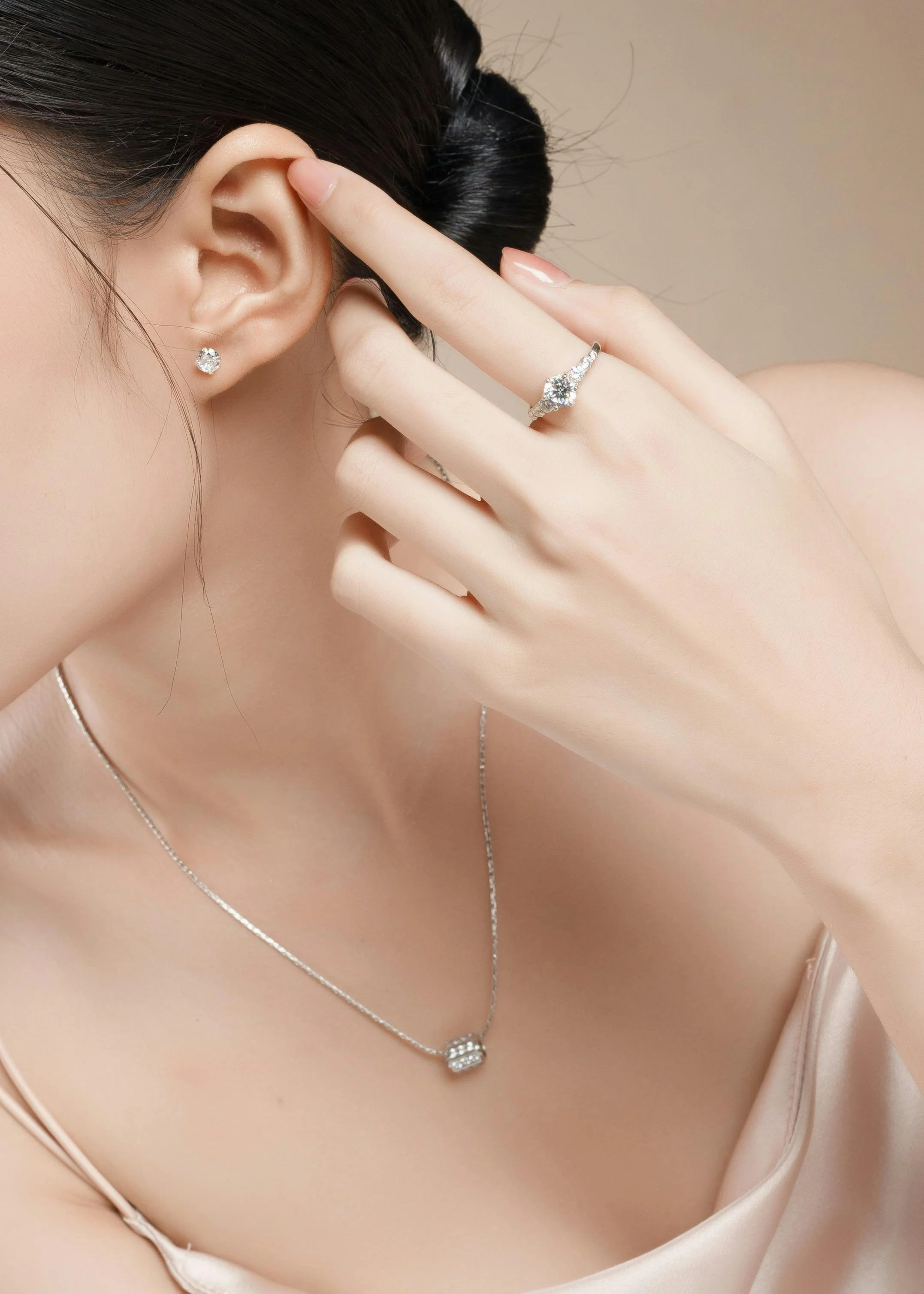 Close-up of a woman with dark hair styled in a bun, wearing diamond earrings, a ring, a necklace, and a satin top, with her hand partially covering her face.