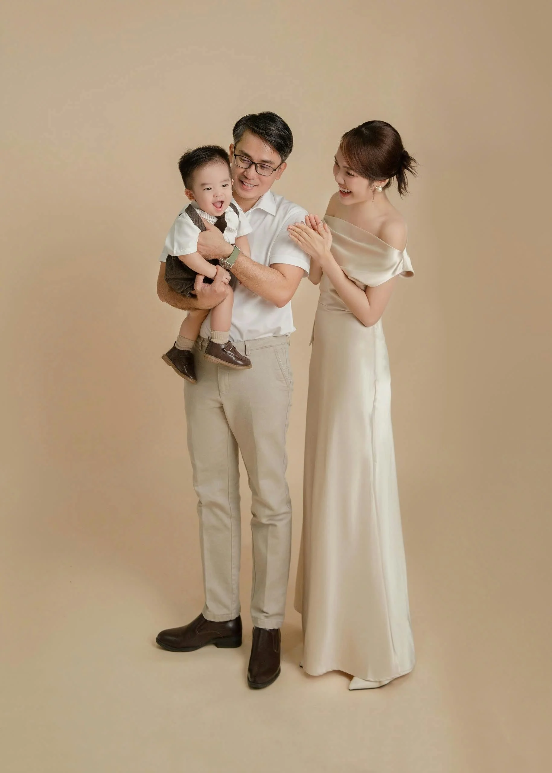 A happy family with a man, woman, and young boy standing together against a beige background, smiling and engaging with each other.