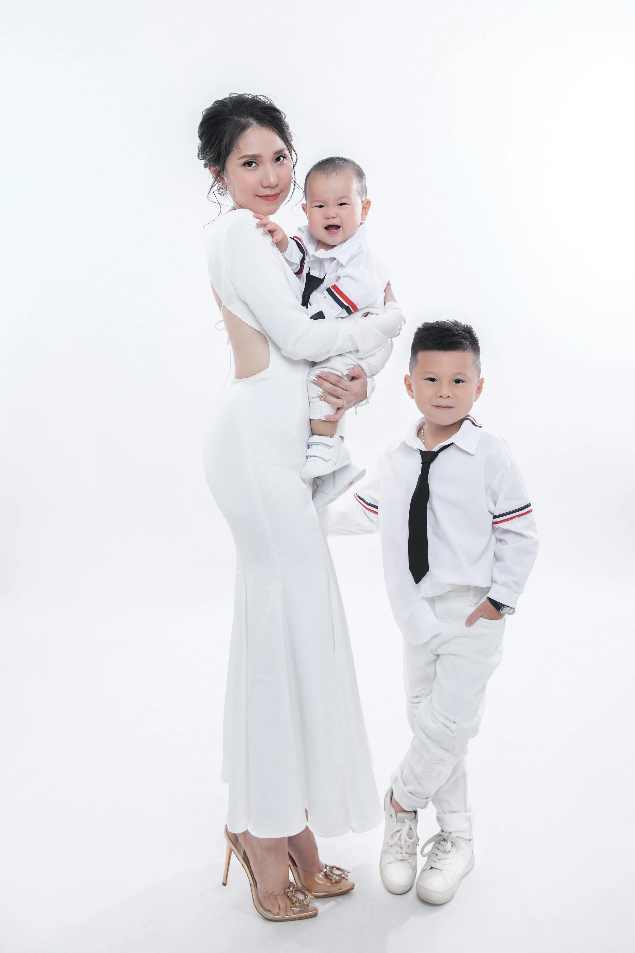 A woman in a white dress holding a crying baby boy, and a boy in a white shirt with a black tie, all standing against a white background.
