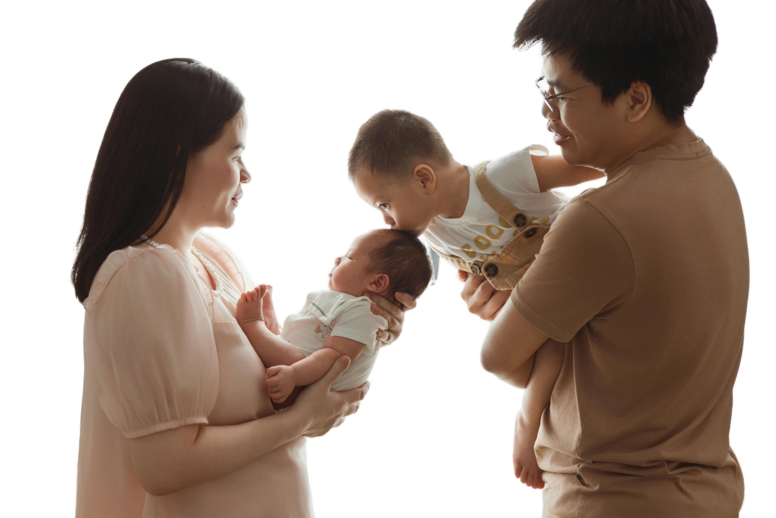 A family of four holding their newborn and young children in front of a white background, facing each other, with the mother and father smiling and the children leaning in toward the babies.