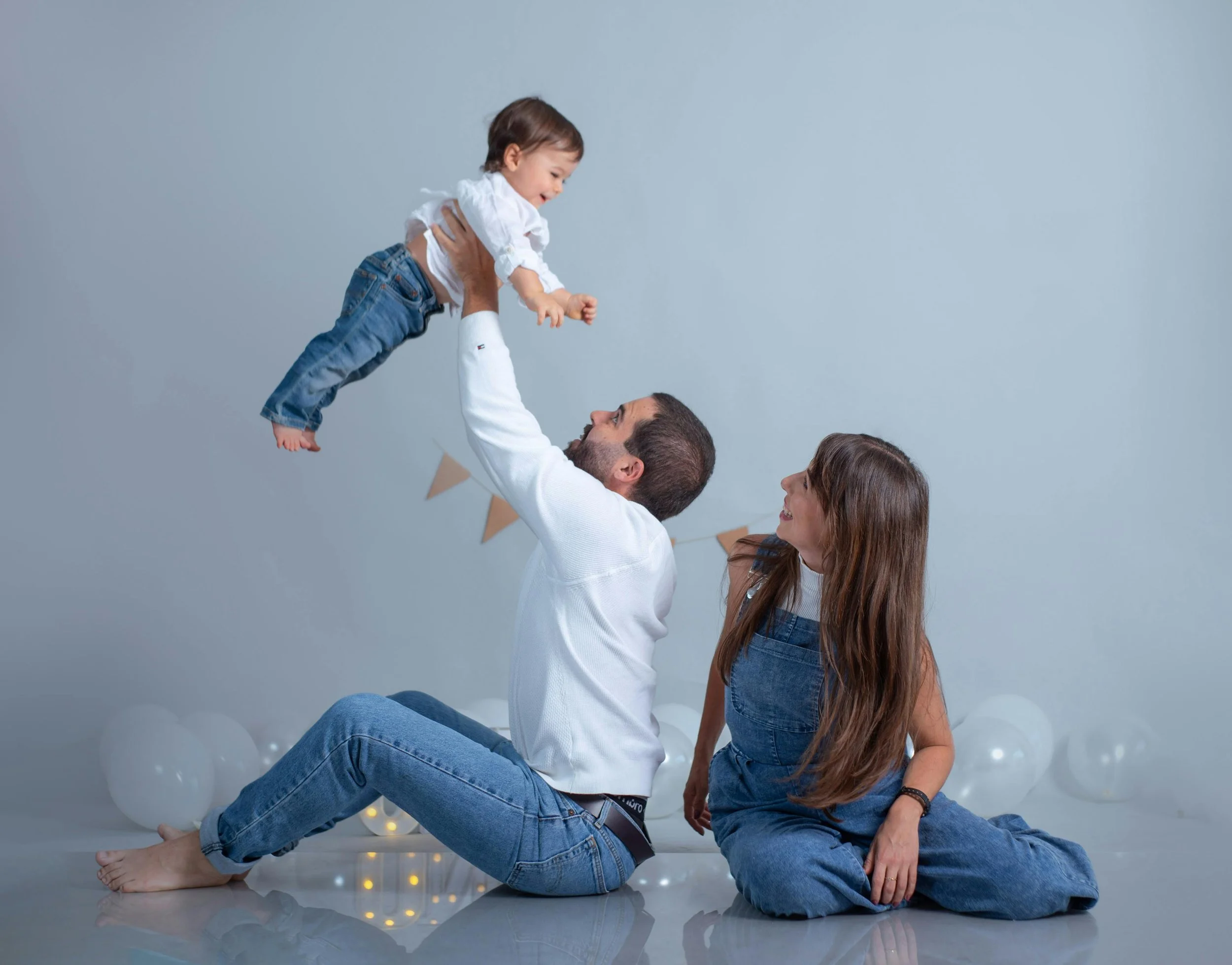 A family of three celebrating together, with a man sitting on the floor lifting a young girl into the air, while a woman sitting nearby watches and smiles, with white balloons and string lights in the background.