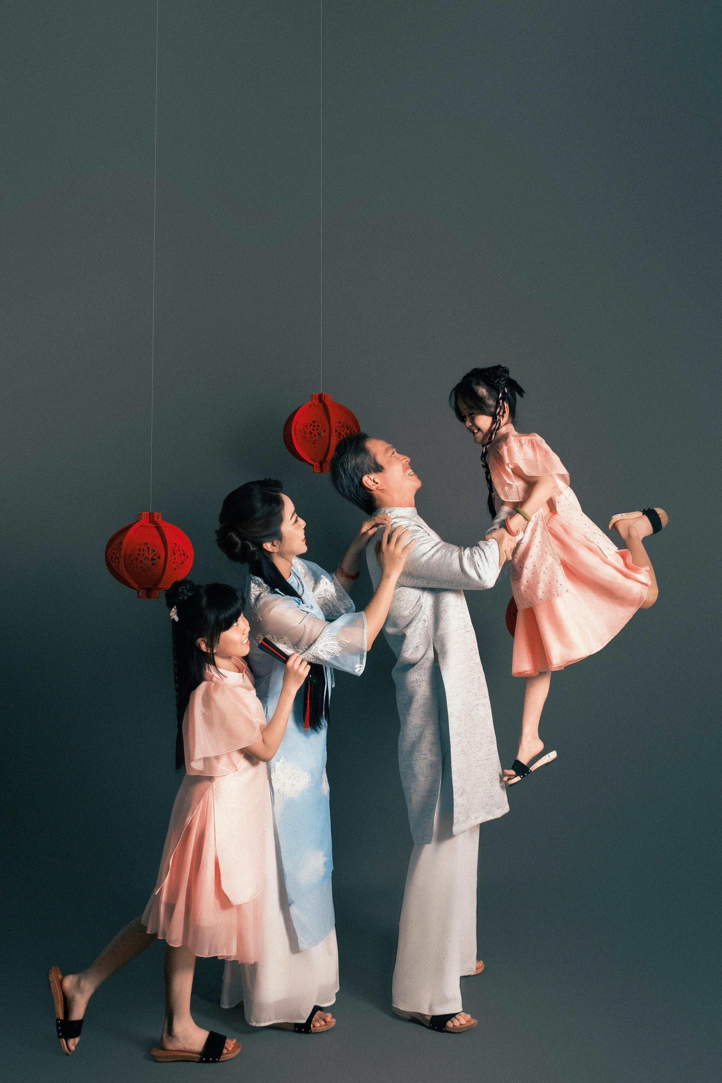 A family of five, consisting of adults and children, enjoying a joyful moment together while holding a young girl in the air. The family is dressed in traditional Asian attire, with red lantern decorations hanging in the background.