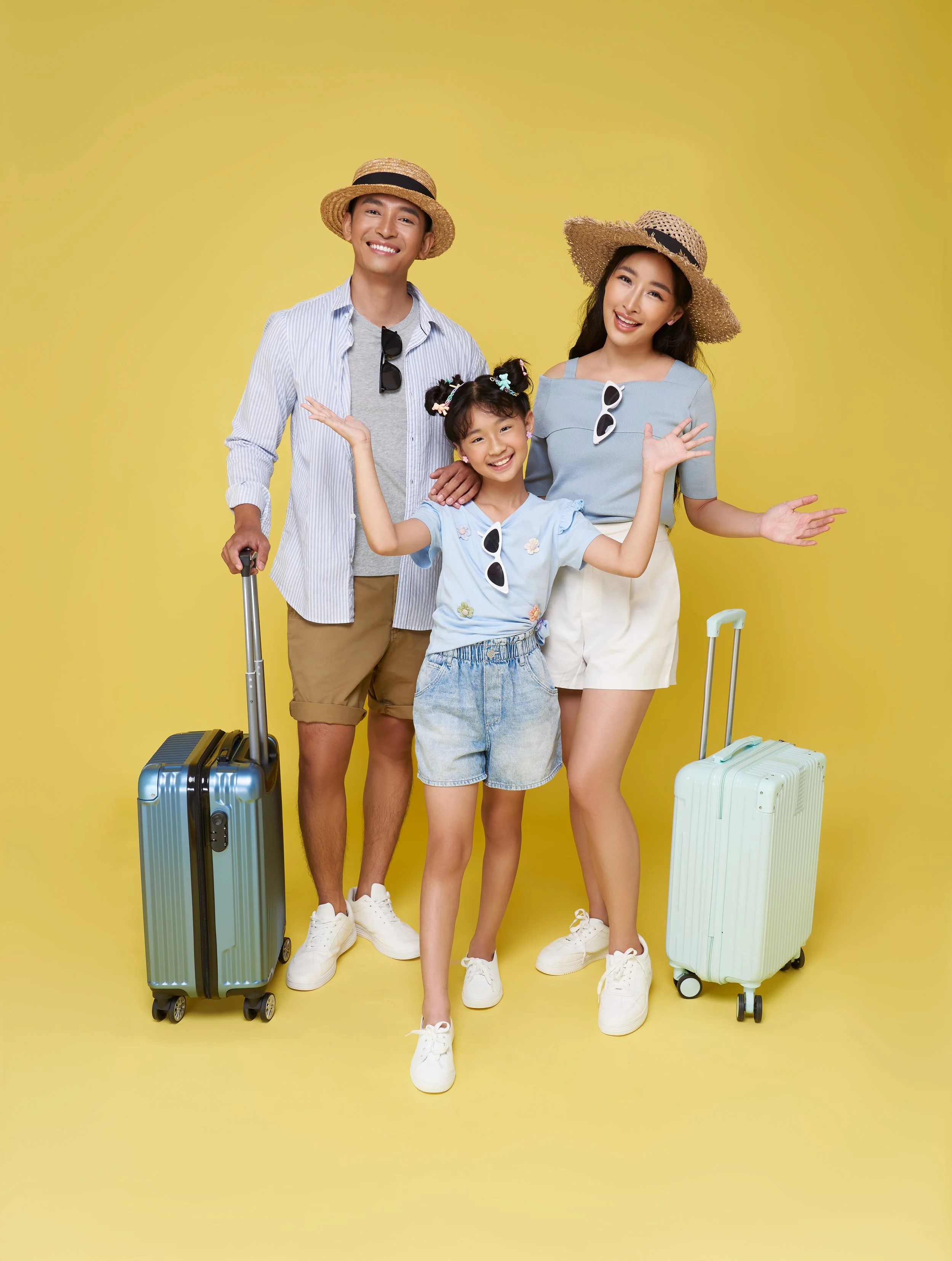 Family of three, two women and one girl, standing with luggage against a yellow background, smiling and wearing summer clothes and hats.