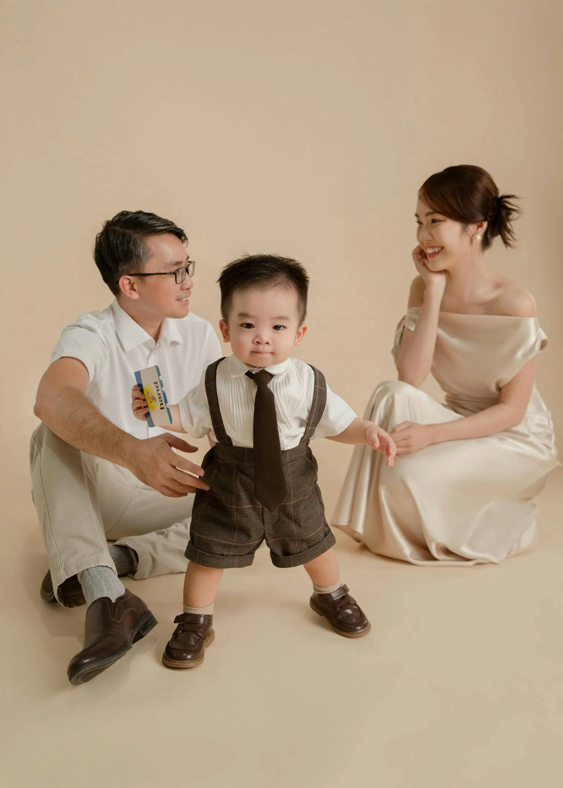 A family of three poses together in a studio with a beige background. The father, wearing glasses and a white shirt, kneels on the floor holding a card. The young boy, dressed in a white shirt, dark brown shorts, and a tie, stands in front, holding o