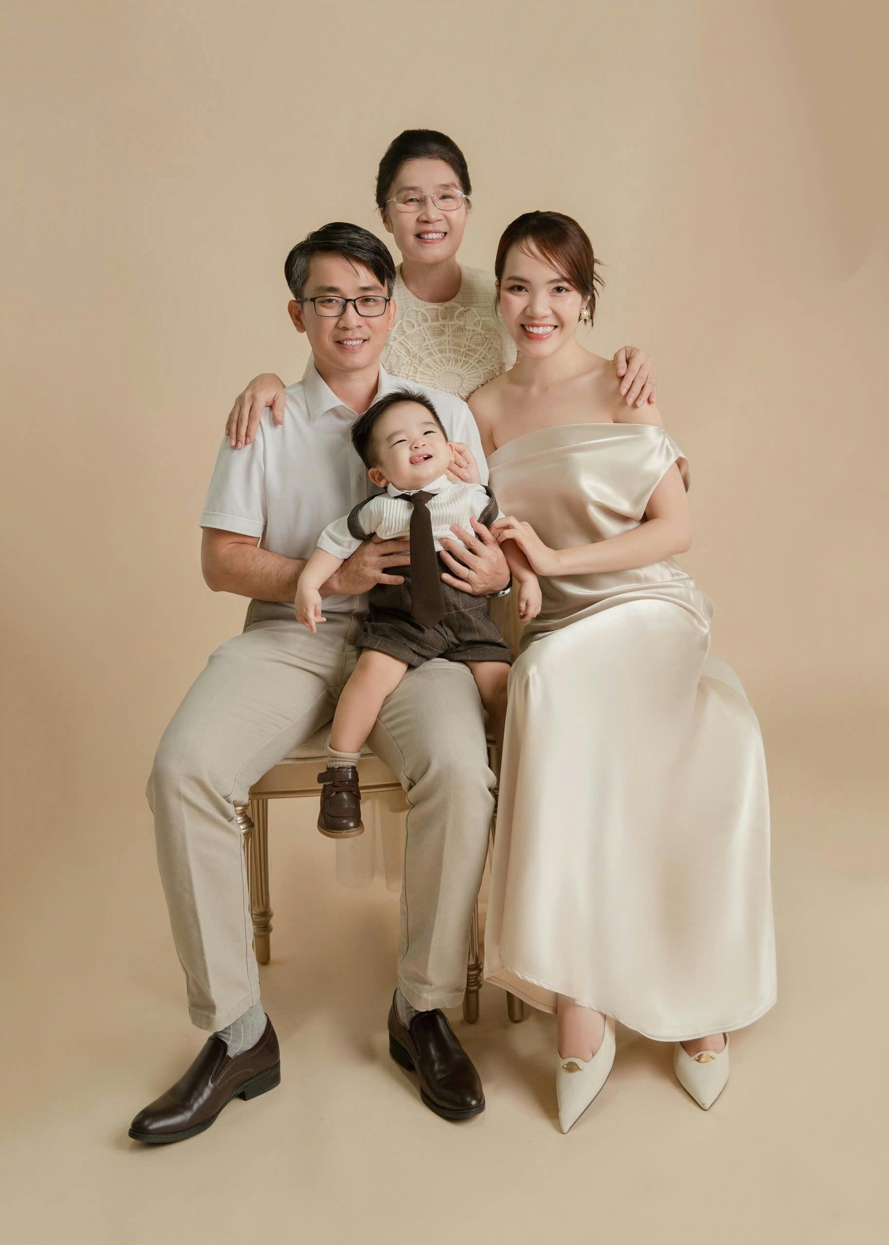 Family portrait of five people, including an elderly woman, a man, a woman, a young boy, and a baby, all smiling and dressed in semi-formal attire, against a plain beige background.