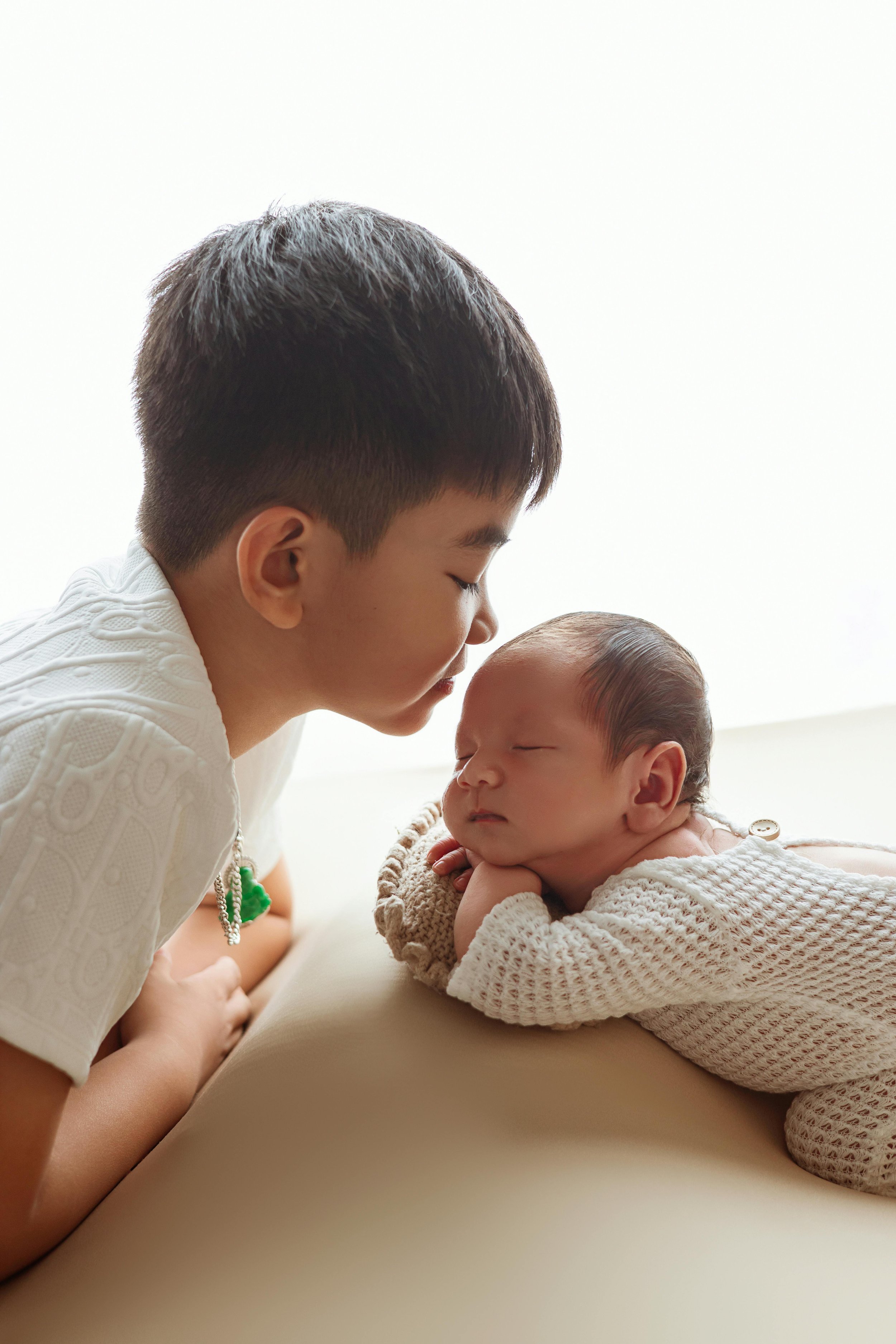 A young boy with dark hair leans close to a sleeping newborn baby with closed eyes, both lying on a soft surface against a bright background.